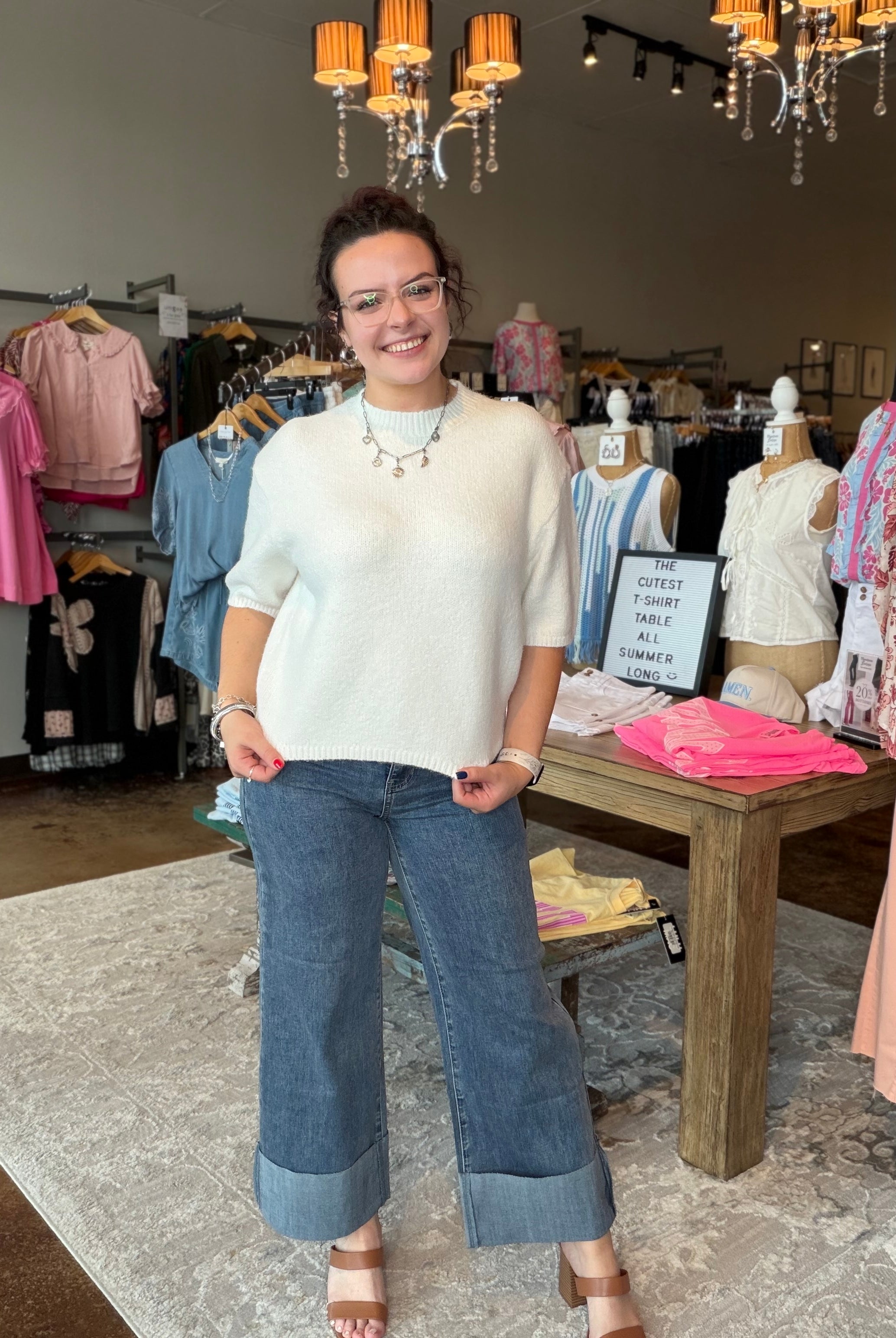 A woman stands in a clothing boutique wearing a cream knit short-sleeve sweater and wide-leg cuffed jeans. The boutique interior features clothing racks, mannequins, a wooden table with folded garments, and hanging chandeliers.