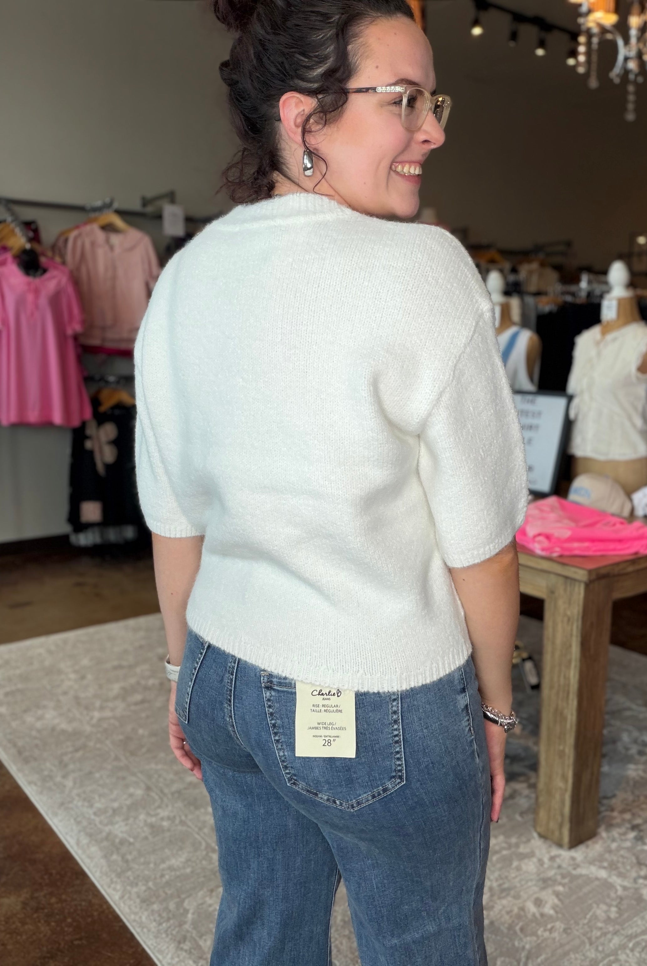 A woman wearing a cream-colored short-sleeve knit sweater and blue jeans, photographed from behind inside a clothing boutique. The back of the jeans features a visible cream brand label that reads 'Cactus'.
