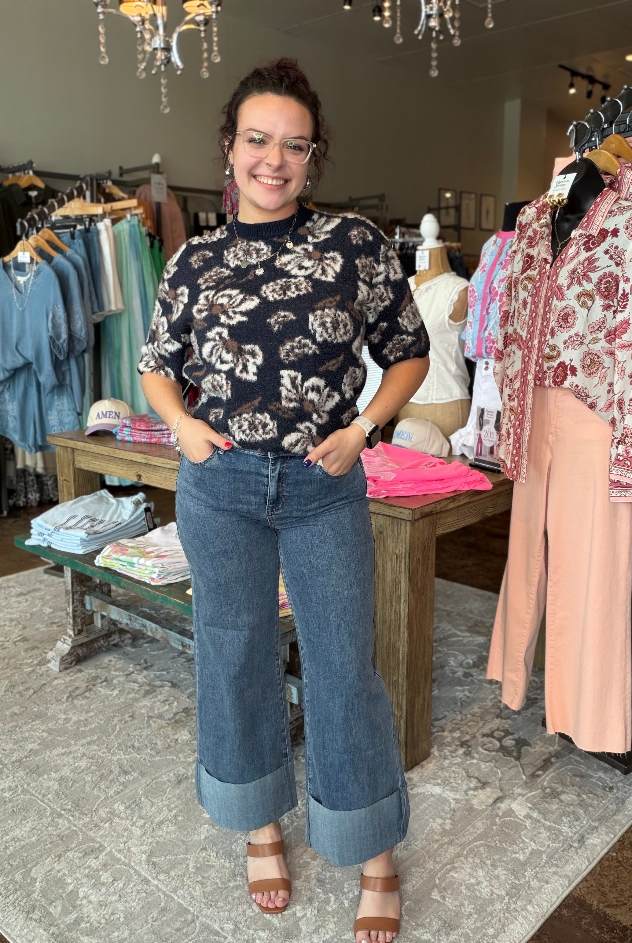 A woman modeling a black short-sleeve floral top and wide-leg blue jeans with cuffed hems, standing inside a clothing boutique.