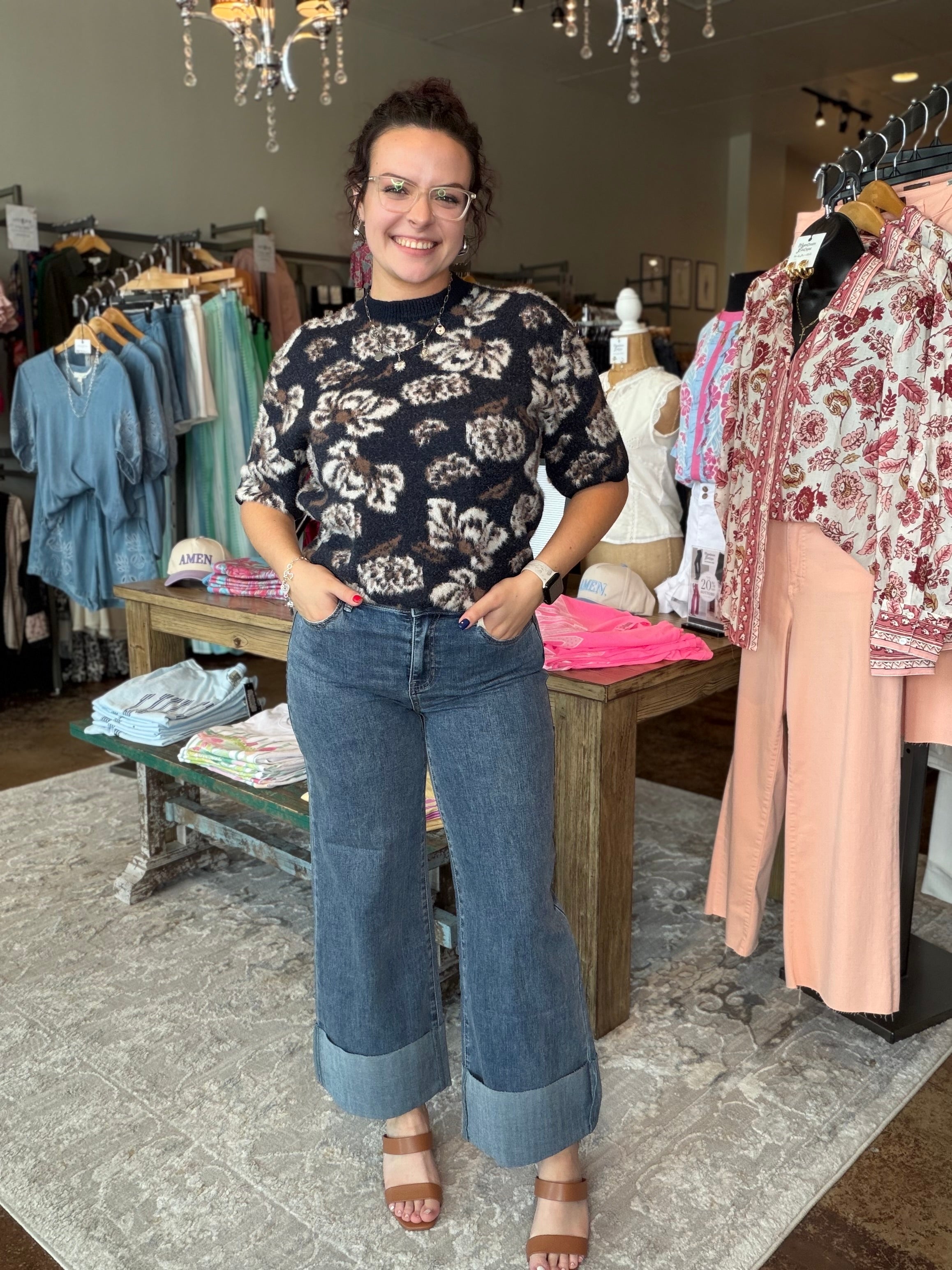 A woman modeling a black short-sleeve floral top and wide-leg blue jeans with cuffed hems, standing inside a clothing boutique.