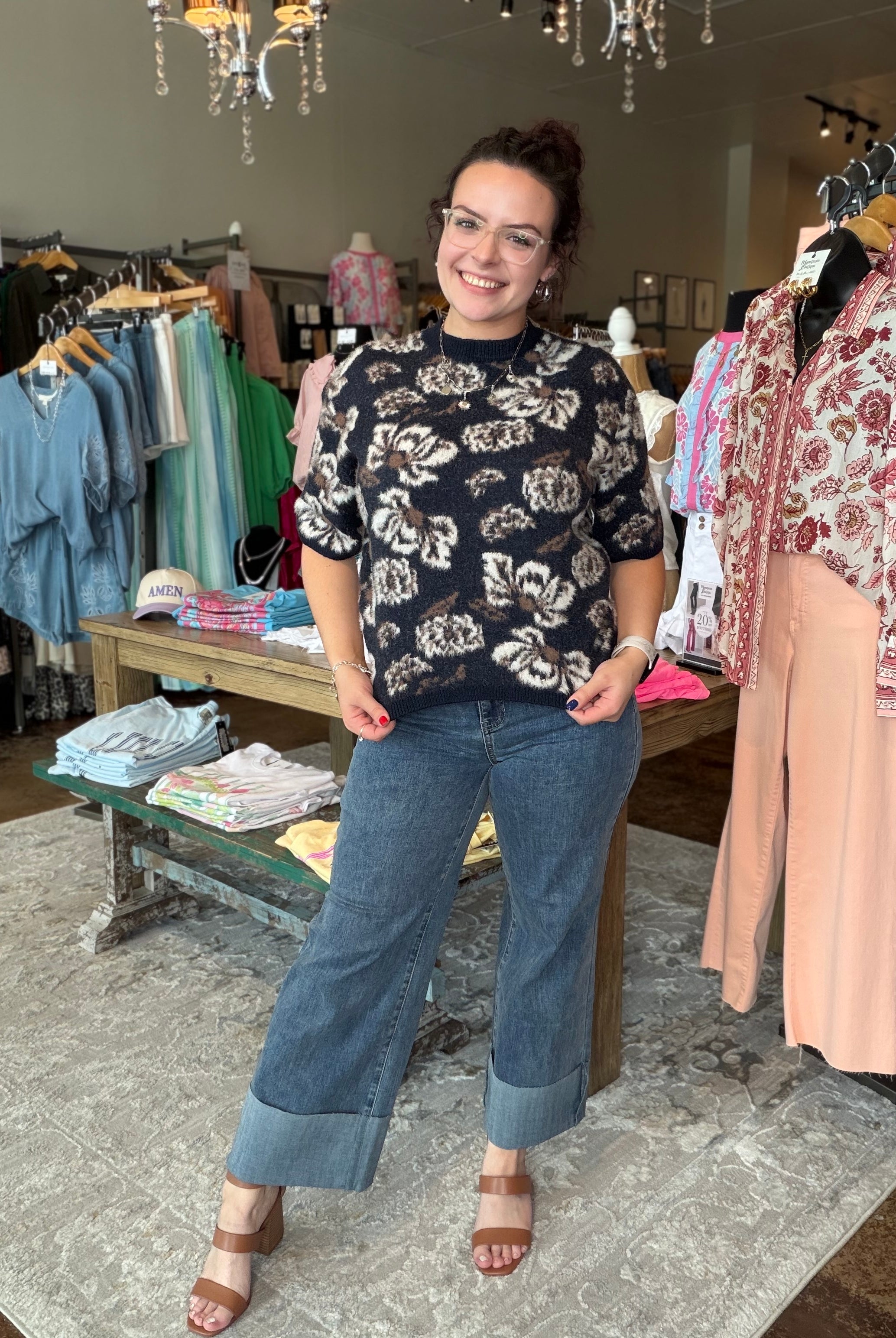 A woman modeling a black short-sleeve floral print top and blue wide-leg jeans with cuffed hems, standing inside a clothing boutique.