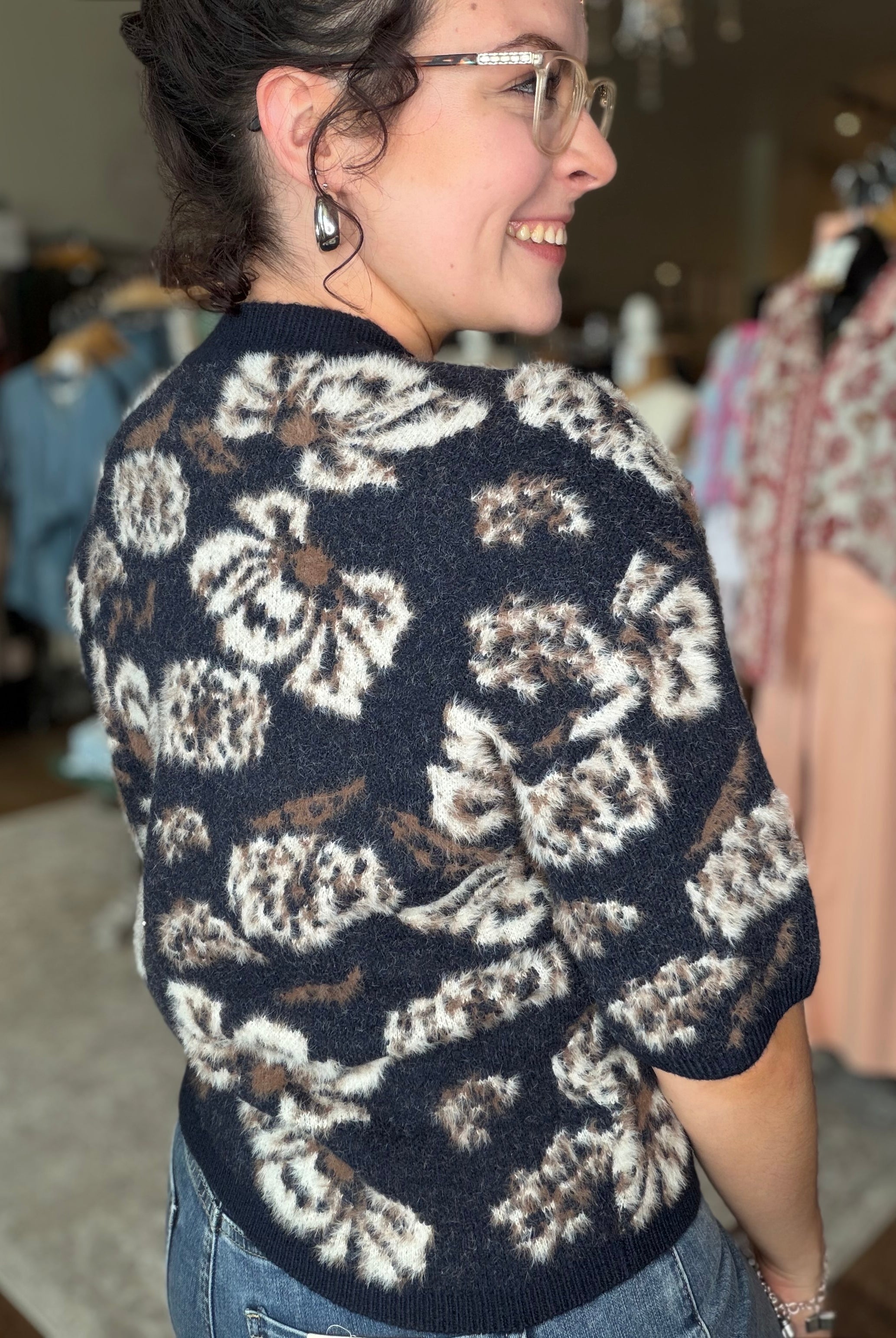 A woman wearing a short-sleeve knit sweater with a large floral pattern in cream, brown, and black, photographed in a clothing store.
