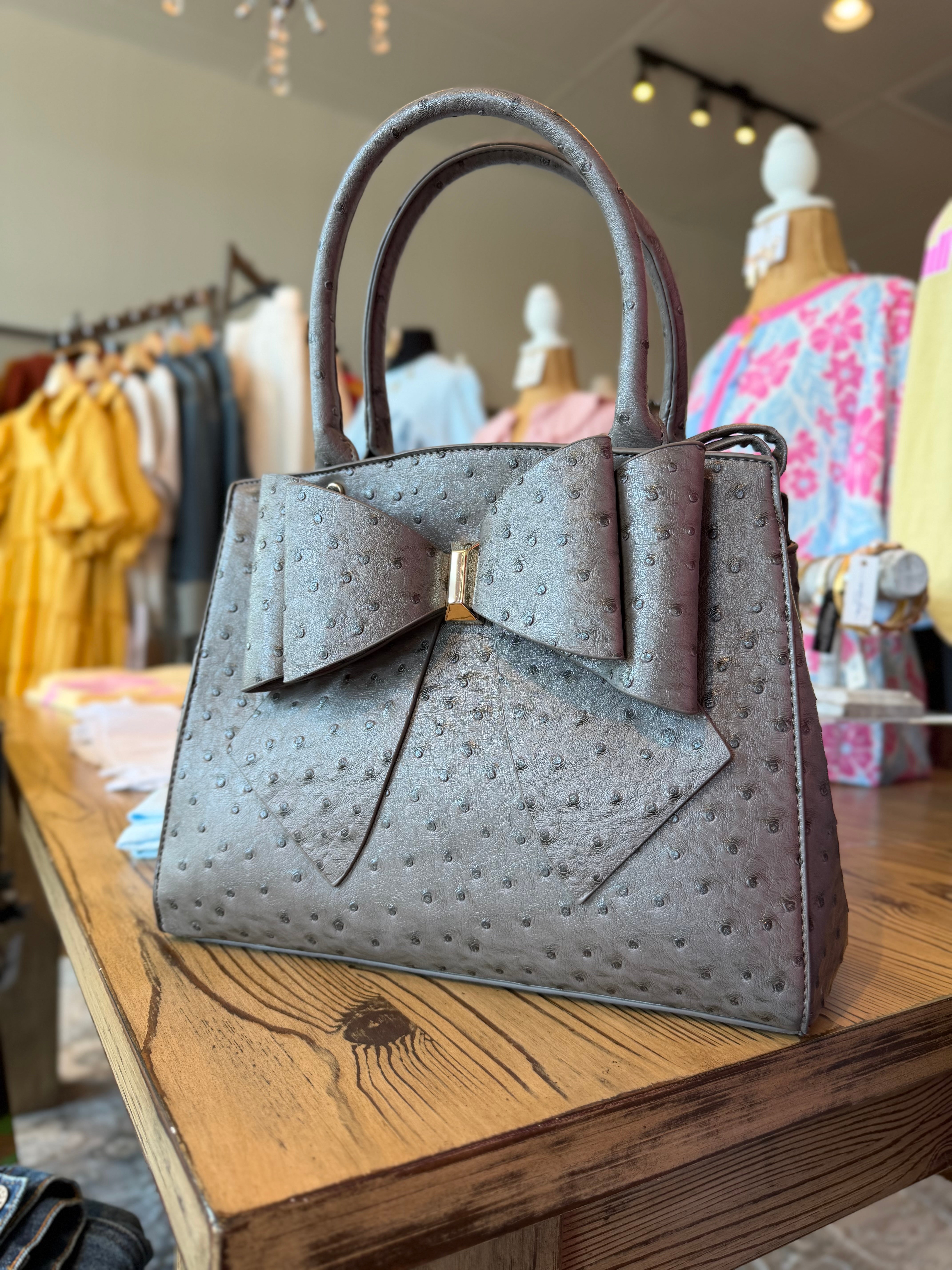 Gray textured handbag with a bow detail on a wooden table in a store setting.