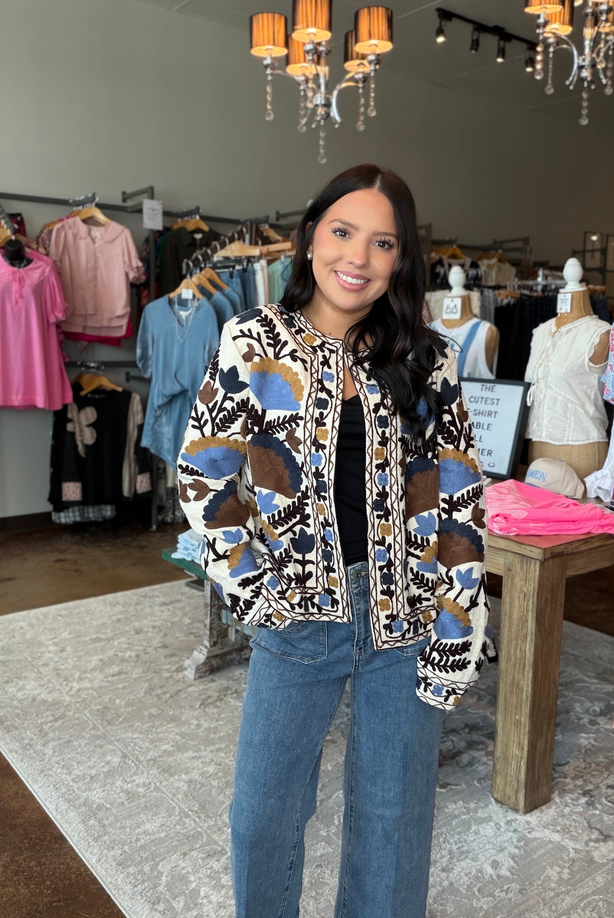 A woman stands inside a clothing boutique wearing a patterned bomber jacket with blue jeans. The jacket features a cream base with abstract geometric patterns in blue, brown, and black.
