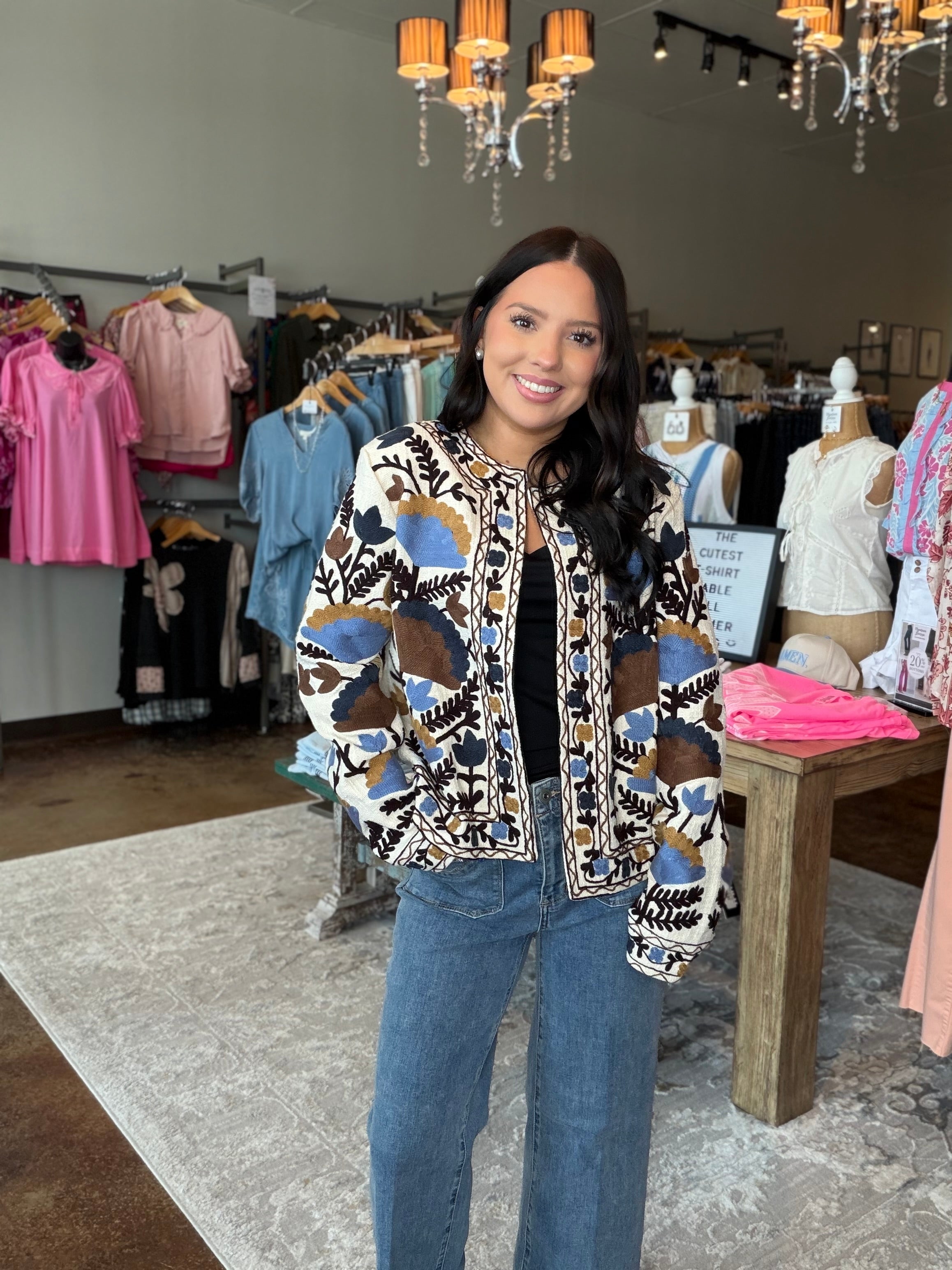 A woman stands inside a clothing boutique wearing a patterned bomber jacket with blue jeans. The jacket features a cream base with abstract geometric patterns in blue, brown, and black.
