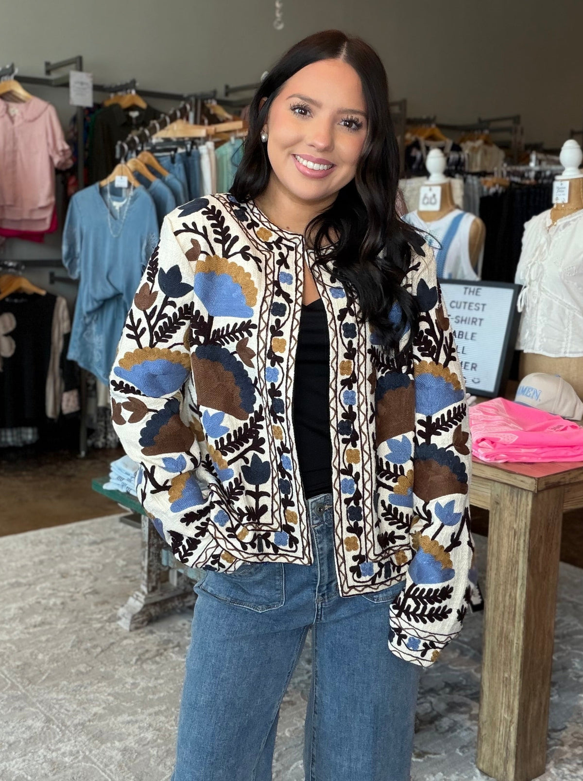 A woman stands inside a clothing boutique wearing a patterned bomber jacket with blue jeans. The jacket features a cream base with abstract geometric patterns in blue, brown, and black.