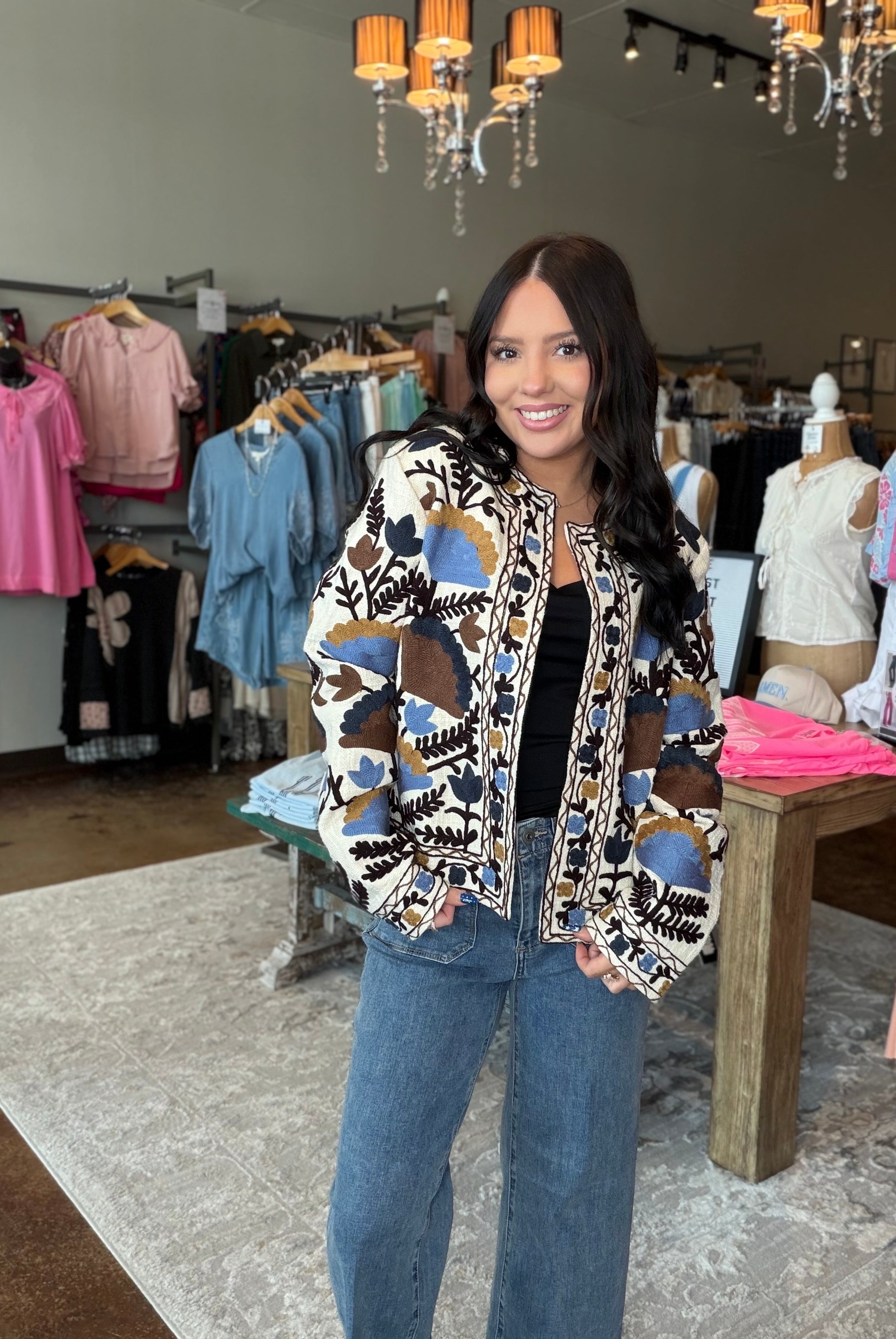 A woman stands inside a clothing boutique wearing a patterned bomber jacket with blue, brown, and beige abstract shapes over a black top and blue jeans.