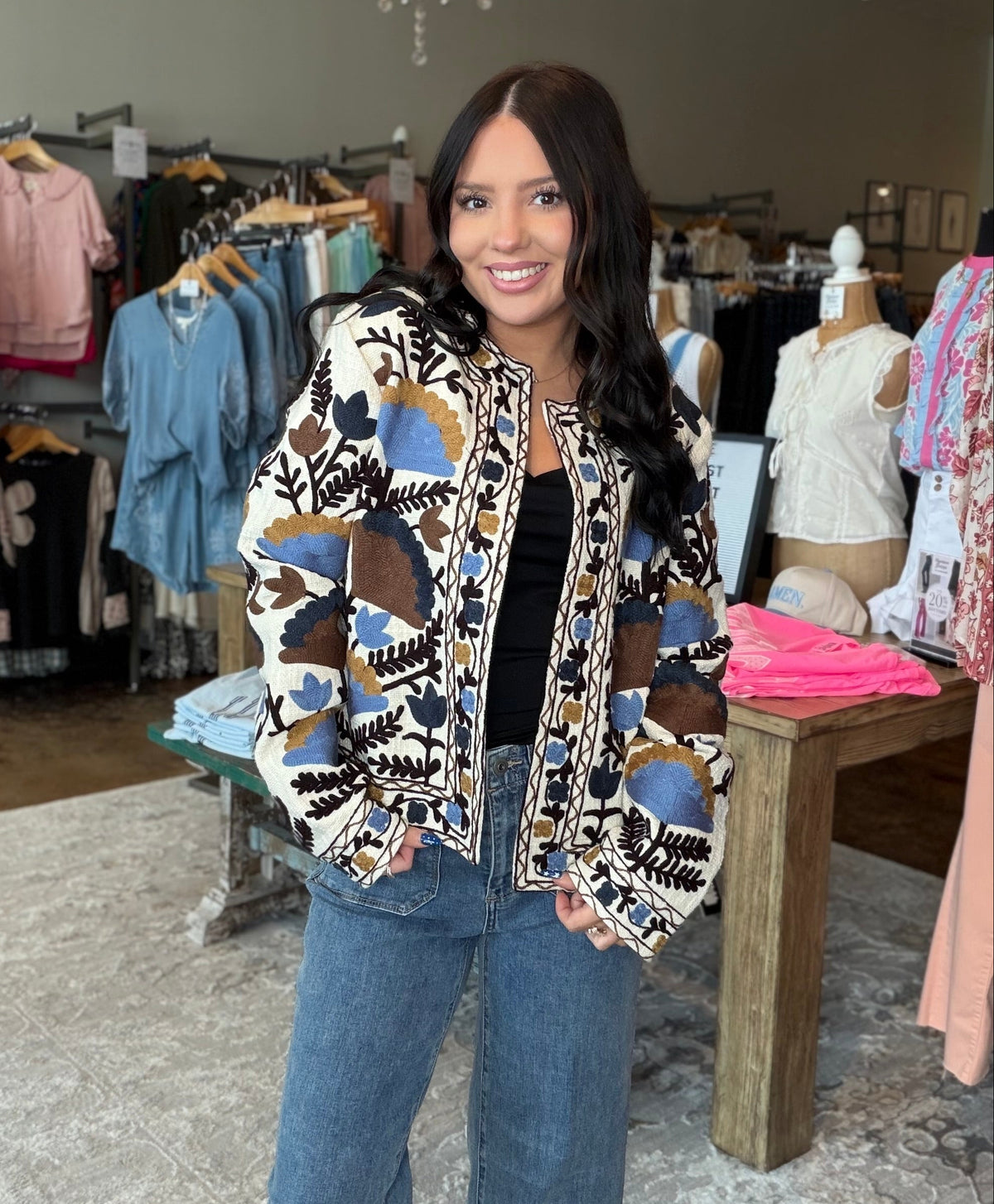 A woman stands inside a clothing boutique wearing a patterned bomber jacket with blue, brown, and beige abstract shapes over a black top and blue jeans.