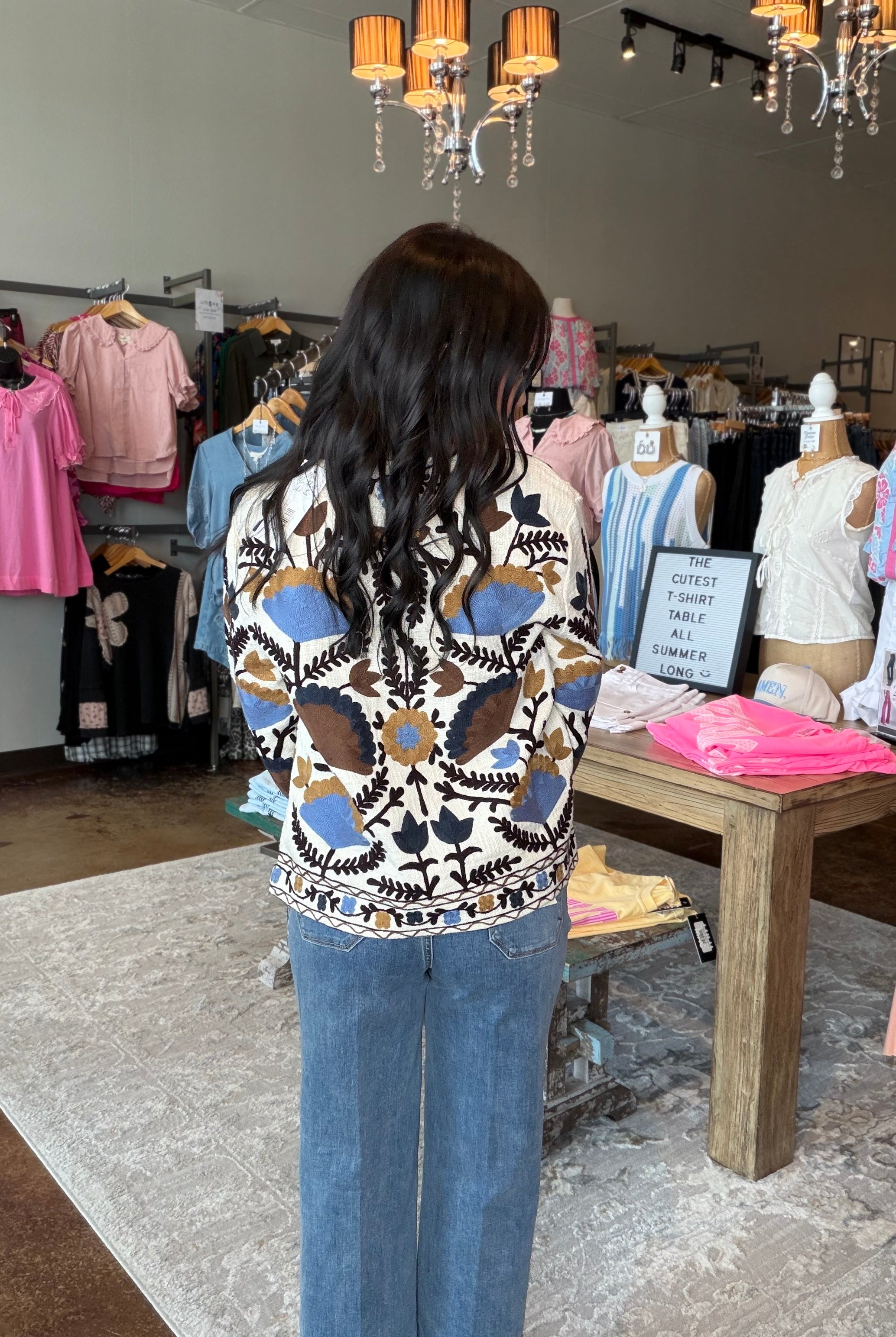 A woman viewed from behind wearing a cream knit sweater with a brown, blue, and black geometric pattern, paired with blue jeans, standing inside a clothing boutique.