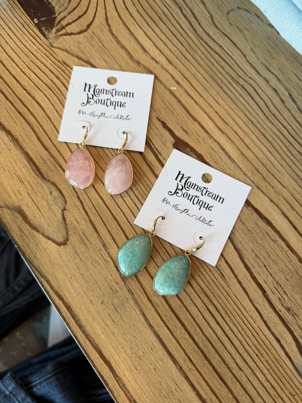 Two pairs of teardrop-shaped drop earrings displayed on white cards on a wooden table. One pair has pale pink stones with gold hooks, and the other has mint green stones with gold hooks.