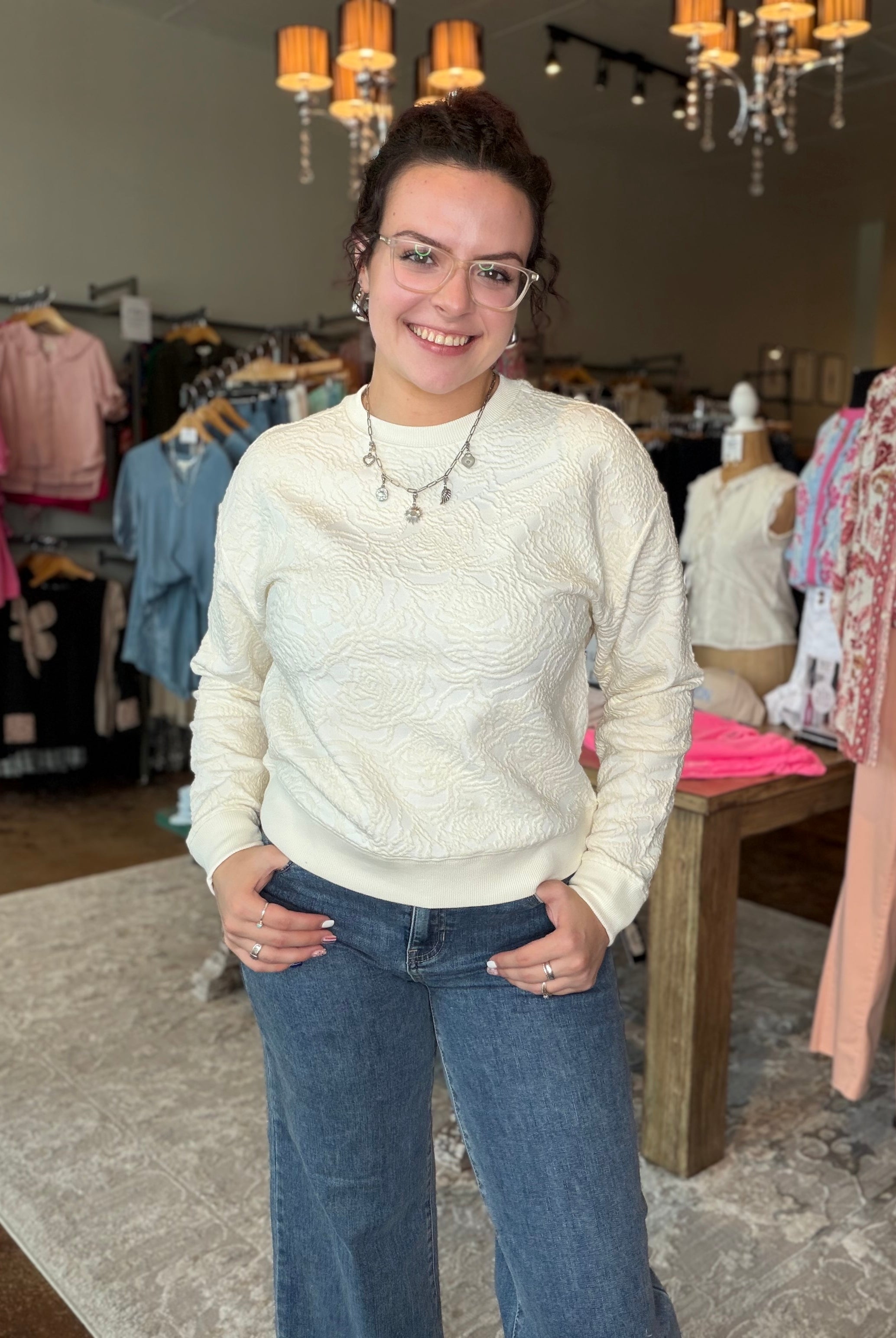 A woman wearing a cream cable-knit long-sleeve sweater and blue jeans stands in a clothing boutique.