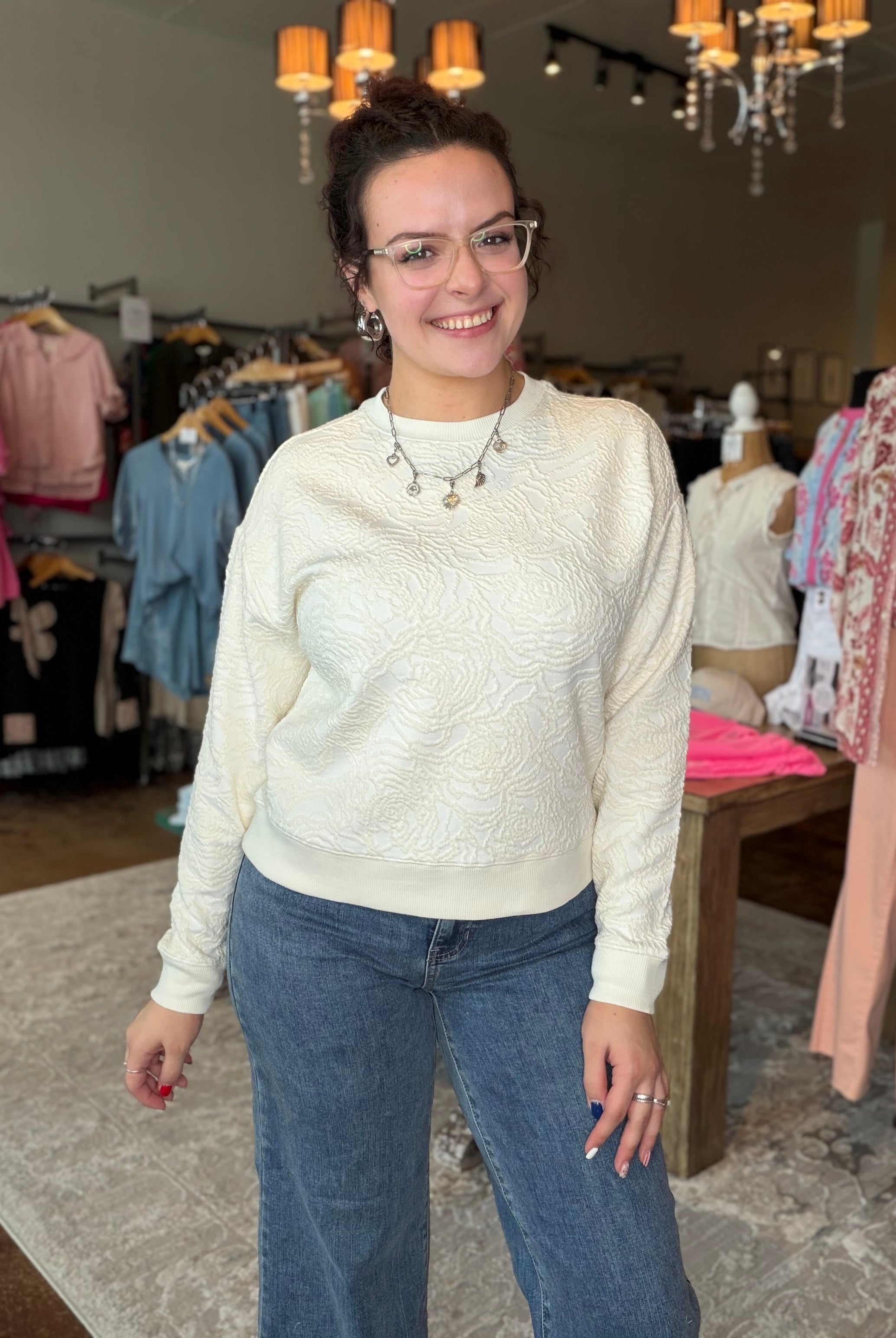 A woman wearing a cream-colored textured knit sweater and blue jeans stands inside a clothing boutique.