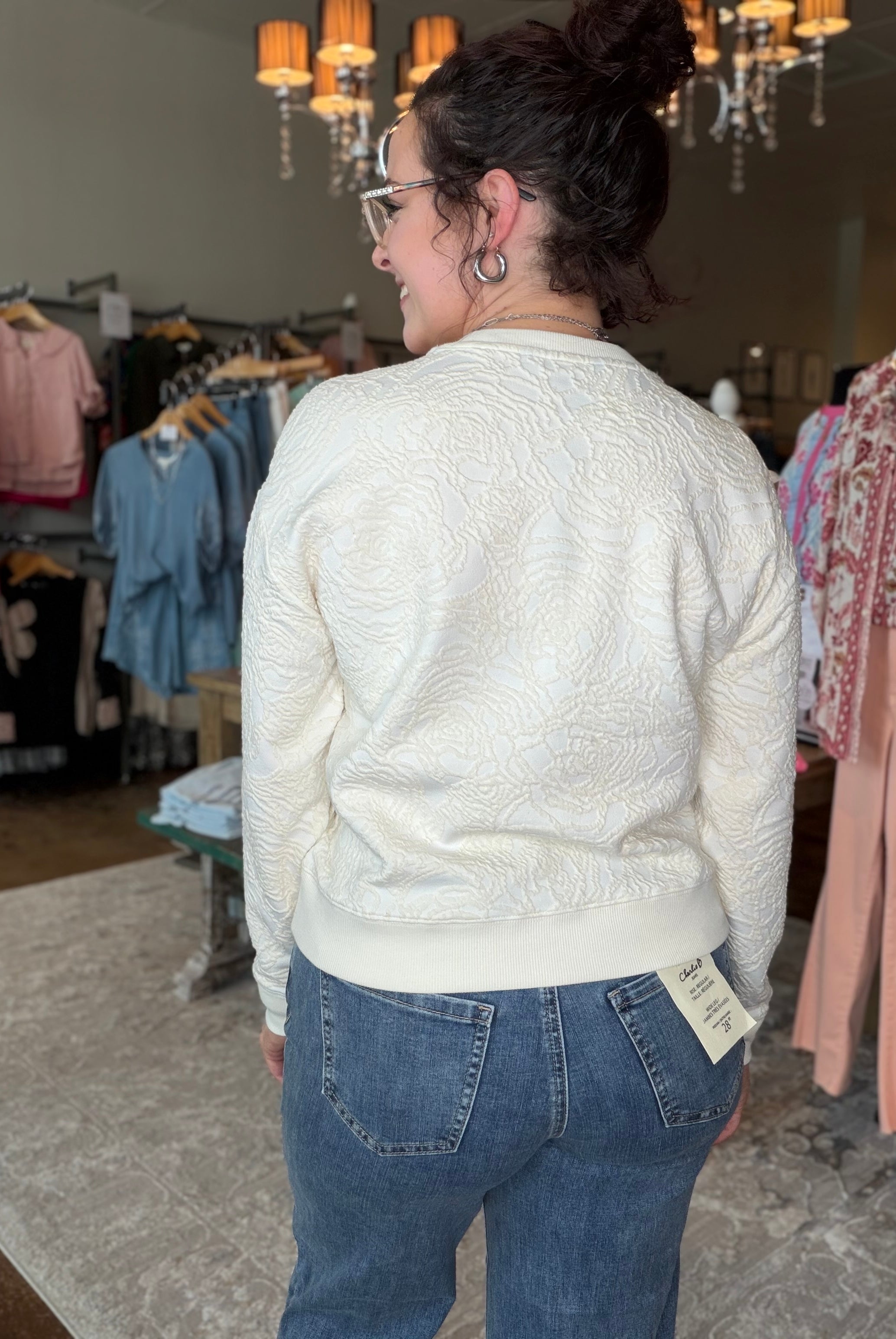 A woman wearing a cream-colored textured knit cardigan and blue jeans, photographed from behind inside a clothing boutique.