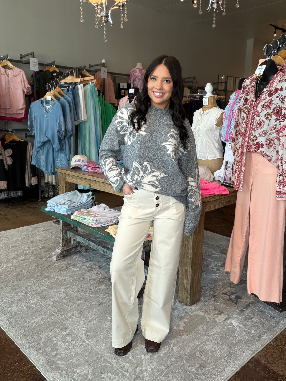A woman stands in a clothing boutique wearing a gray floral knit sweater and cream wide-leg pants. Clothing racks, mannequins, a chandelier, and a patterned rug are visible in the store interior.