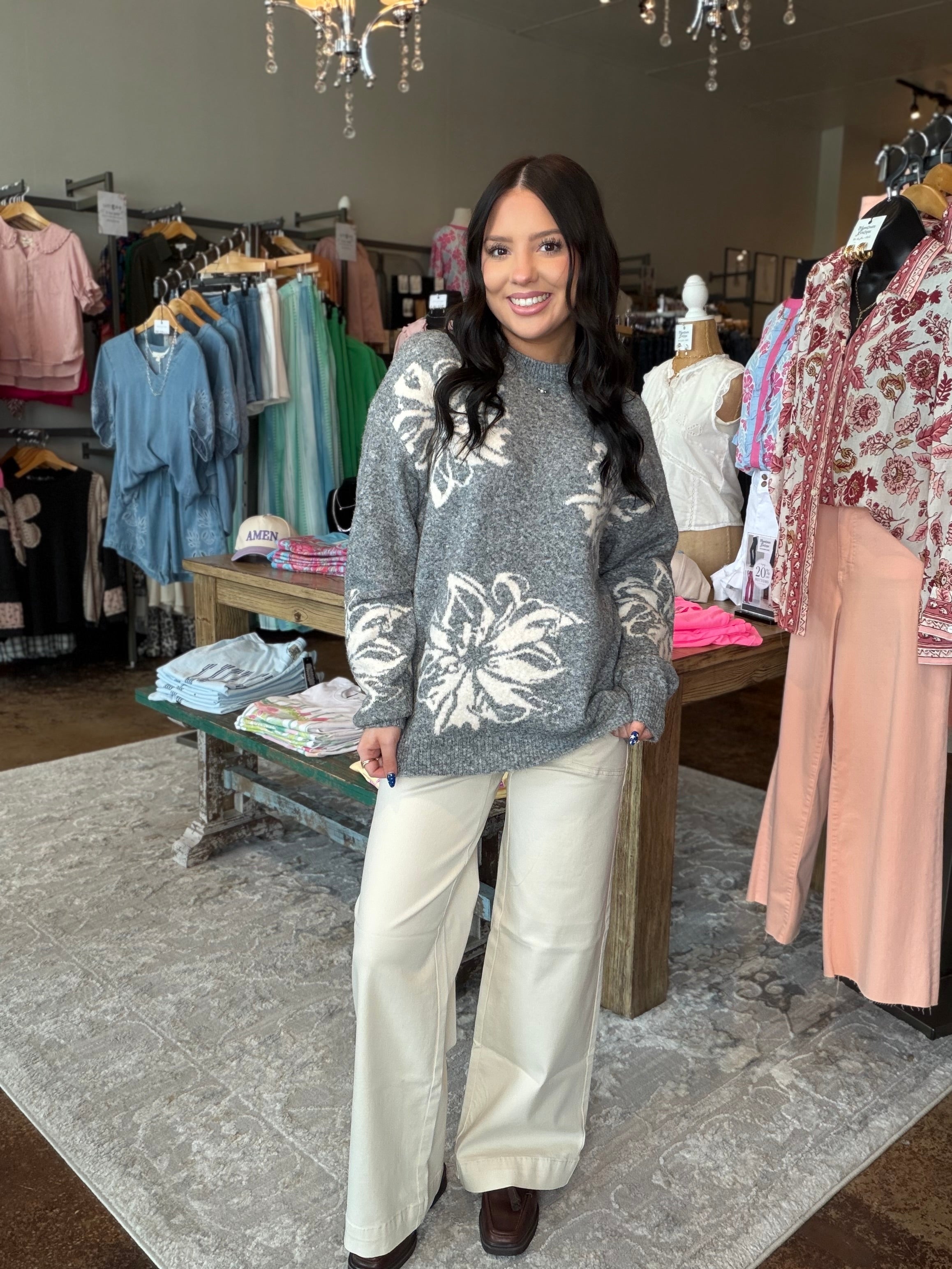 A woman stands in a clothing boutique wearing a gray knit sweater with large white floral patterns and wide-leg cream pants. Clothing racks, mannequins, and store fixtures are visible in the background.