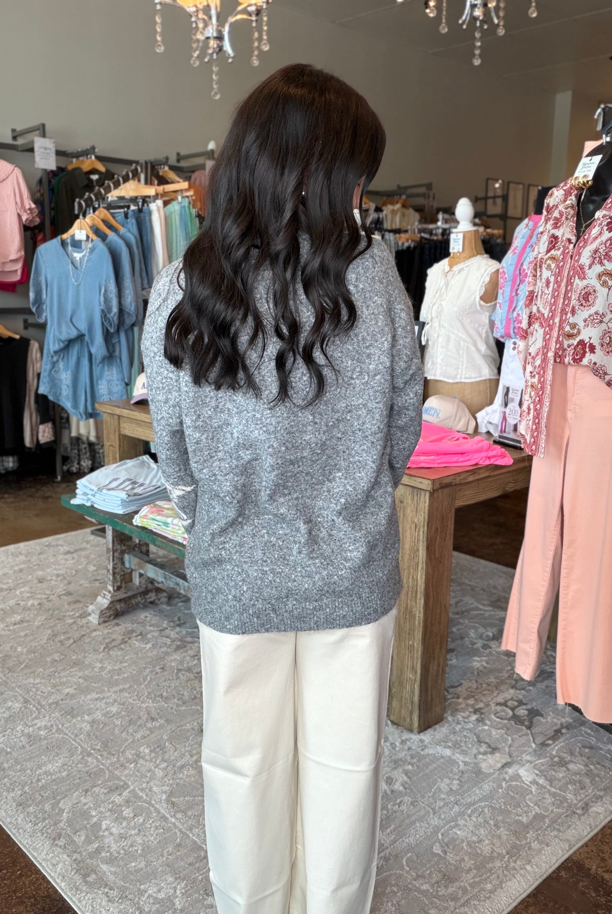 A woman with long, wavy dark hair is seen from behind, wearing a gray knit sweater and cream wide-leg trousers inside a clothing boutique. Clothing racks, a wooden table, and a patterned rug are visible in the background.