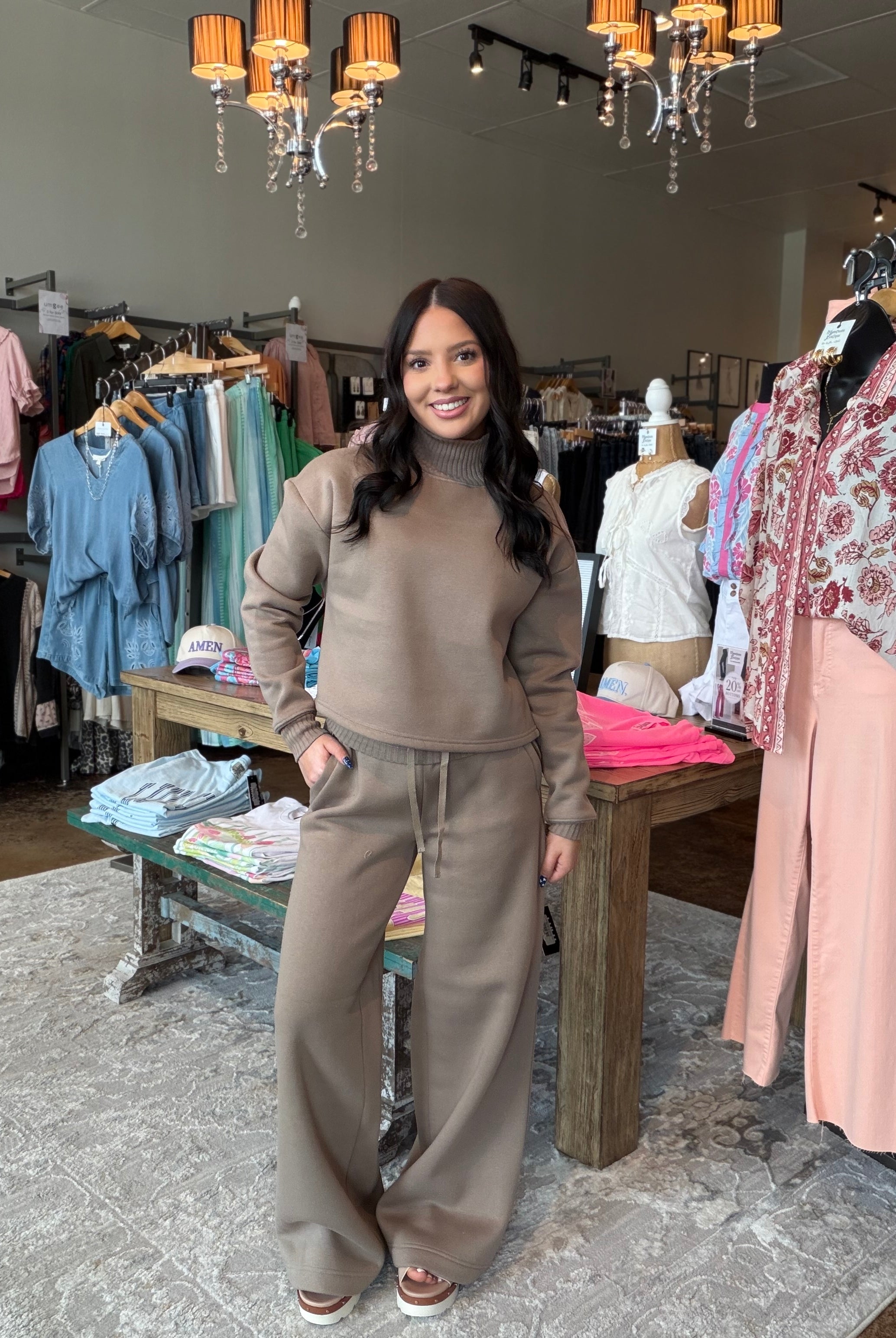 A woman modeling a matching taupe brown lounge set consisting of a turtleneck top and wide-leg sweatpants, standing inside a clothing boutique.