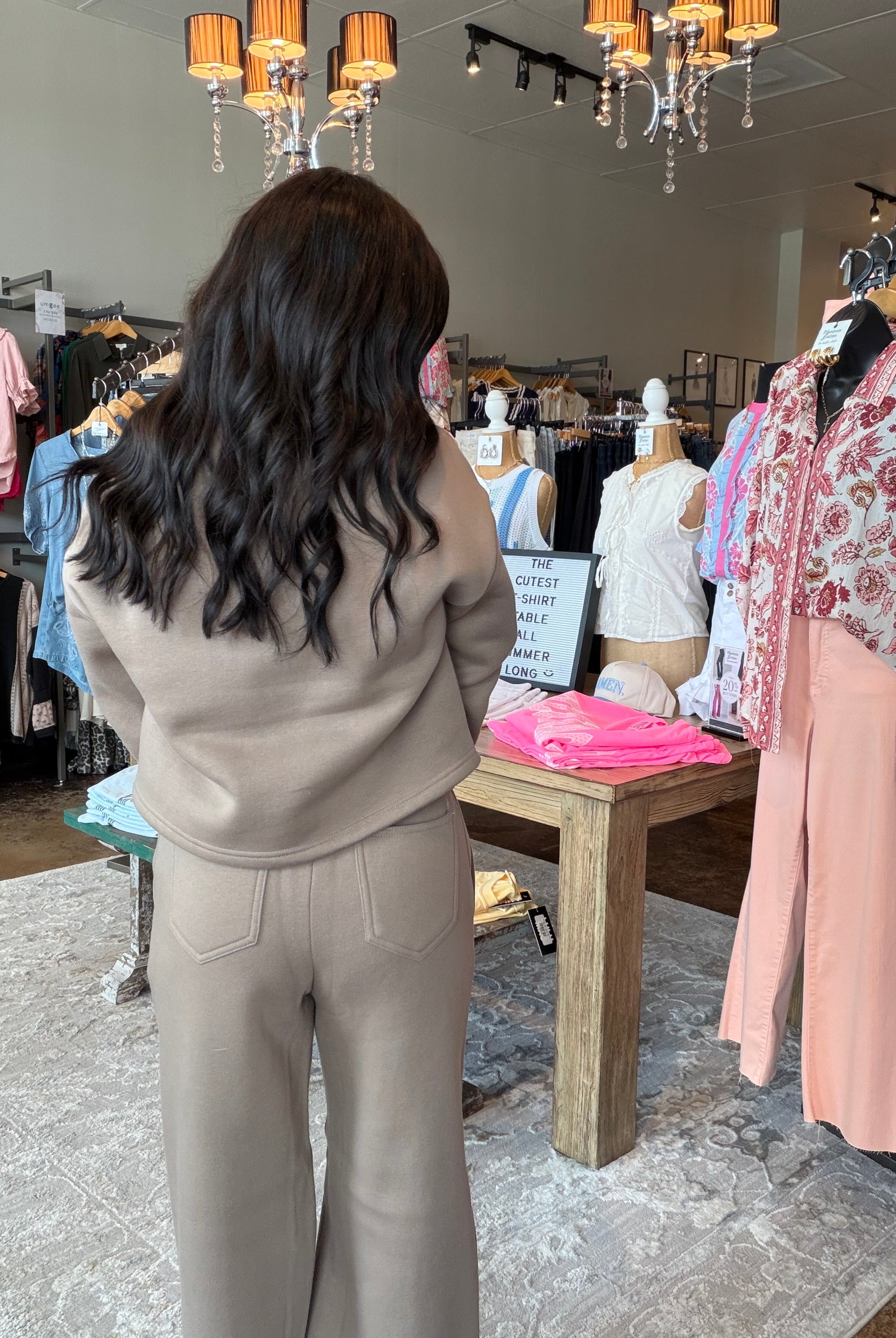A woman with long dark wavy hair stands in a clothing boutique wearing a beige knit matching set consisting of a cropped sweatshirt and wide-leg pants. She is viewed from behind, with racks of colorful clothing, mannequins, and hanging lamps visible in the background.