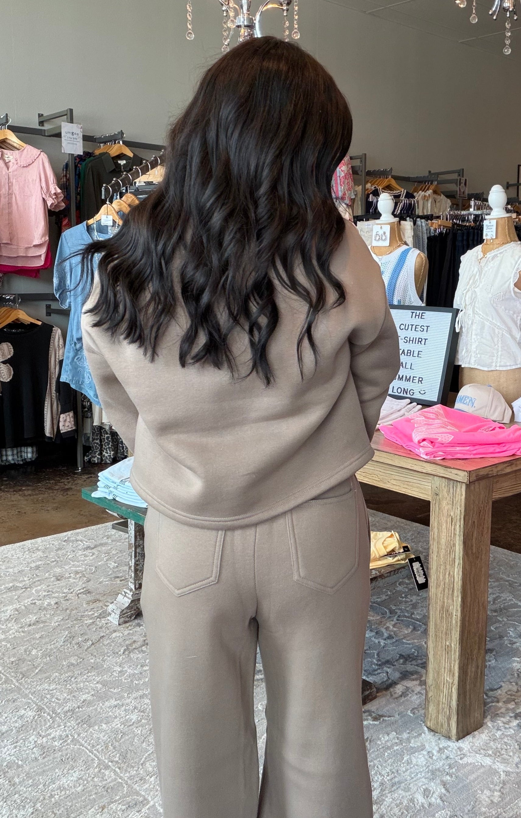 A woman with long dark wavy hair stands in a clothing boutique wearing a beige knit matching set consisting of a cropped sweatshirt and wide-leg pants. She is viewed from behind, with racks of colorful clothing, mannequins, and hanging lamps visible in the background.
