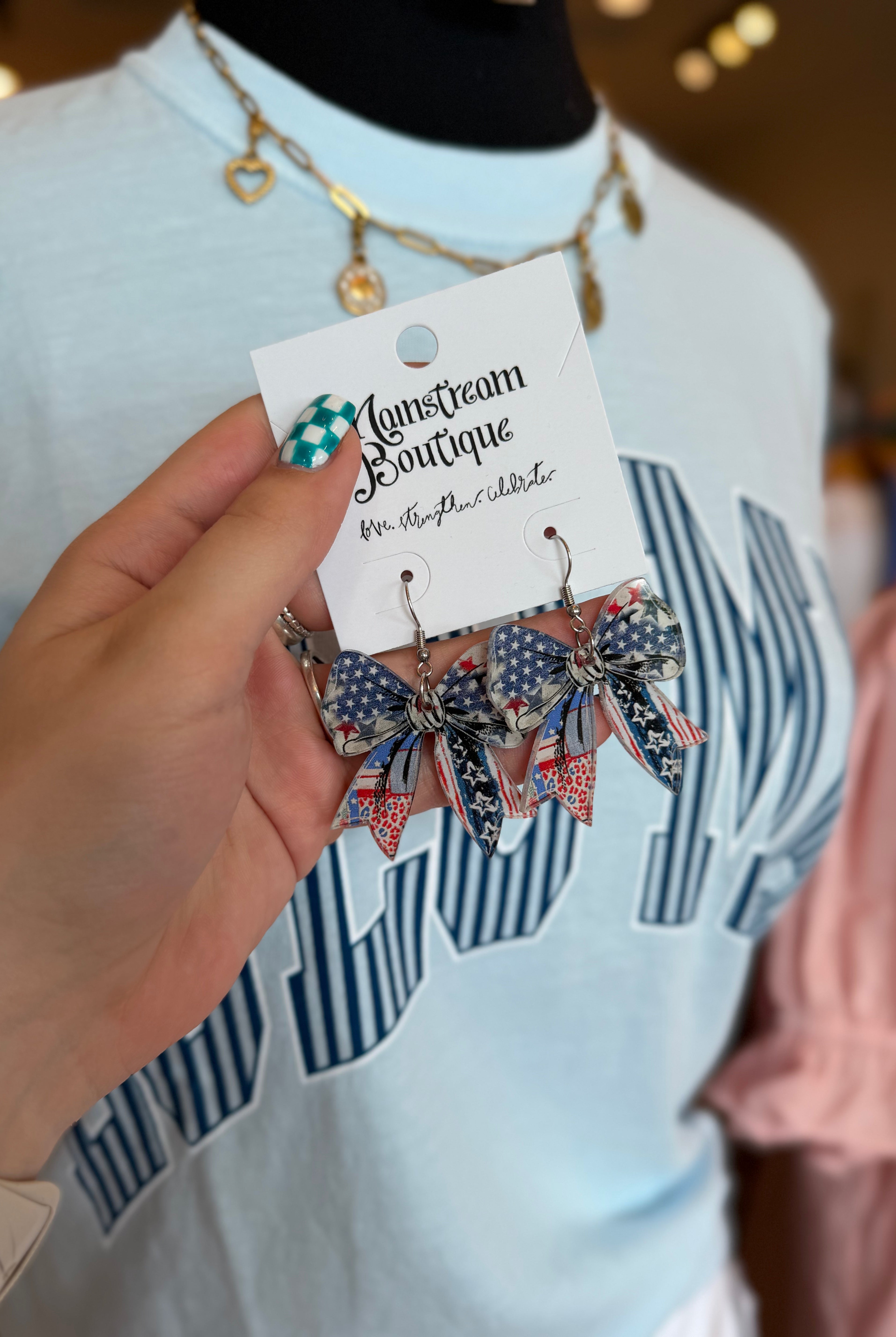 Hand holding a pair of bow-shaped earrings with American flag design on a 'Lansdowne Boutique' card.