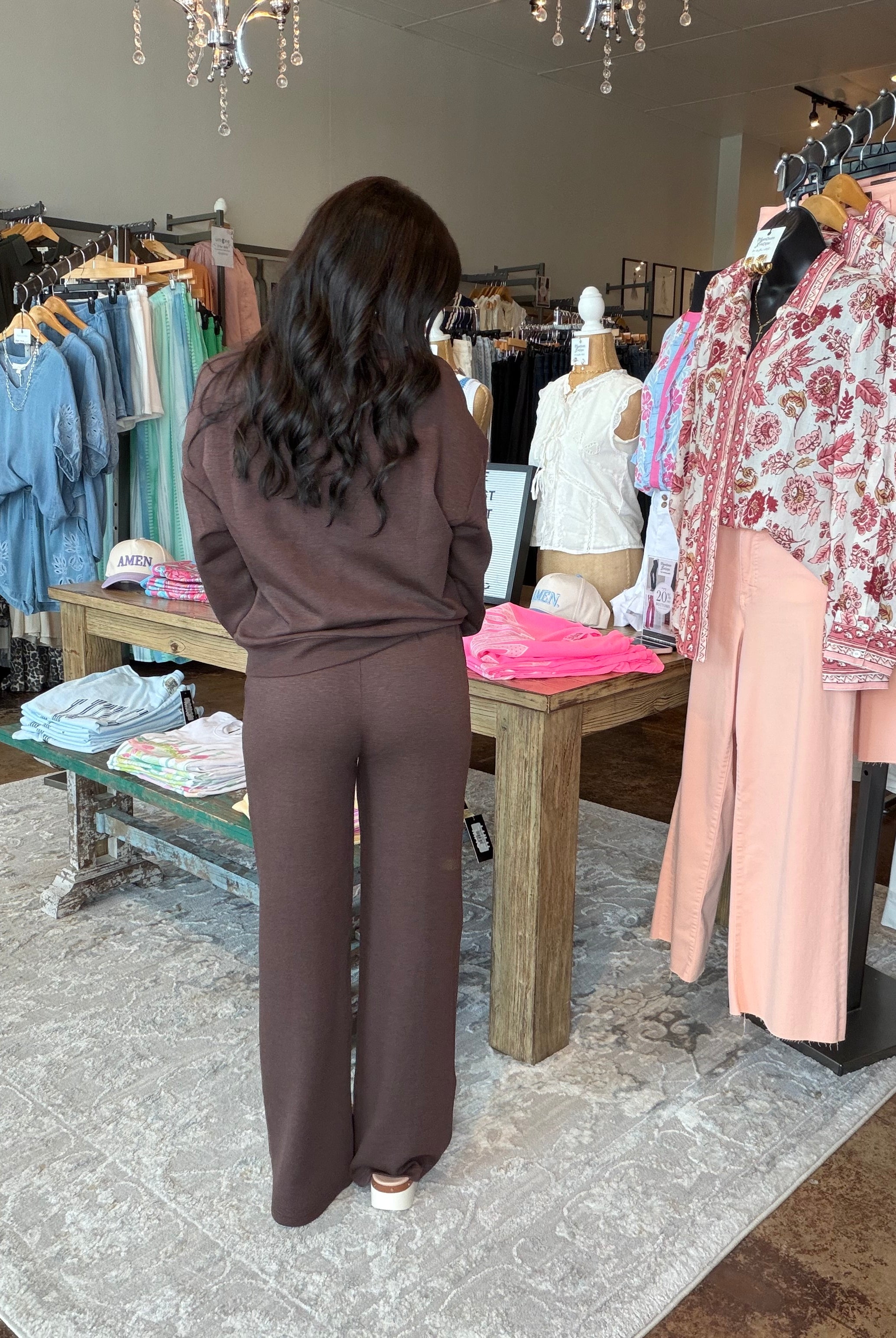 A woman viewed from behind wearing a matching dark brown lounge set consisting of a long-sleeve top and wide-leg pants, standing inside a clothing boutique.