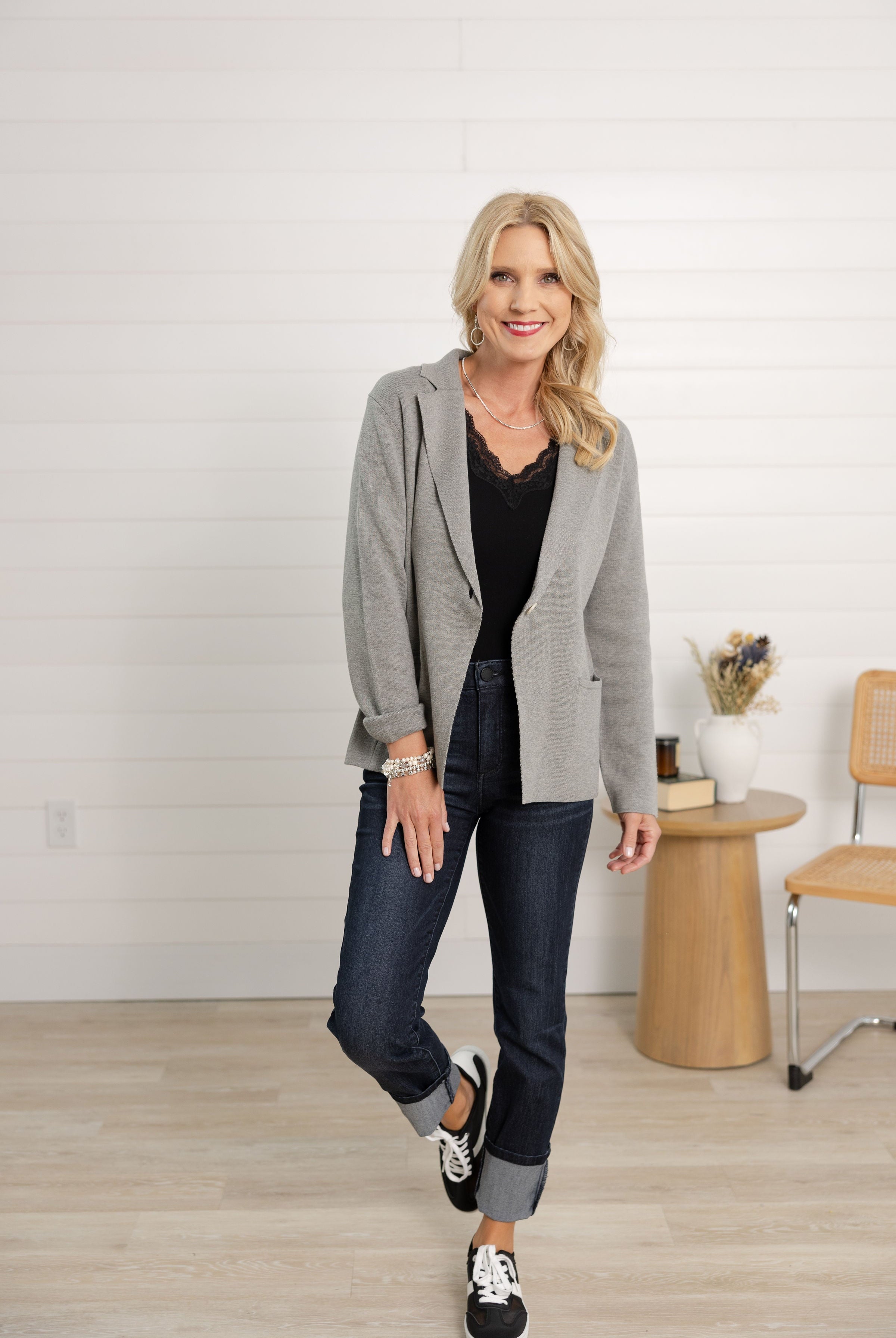 Woman wearing a gray blazer, black top, and jeans in a room with a wooden table and chair.