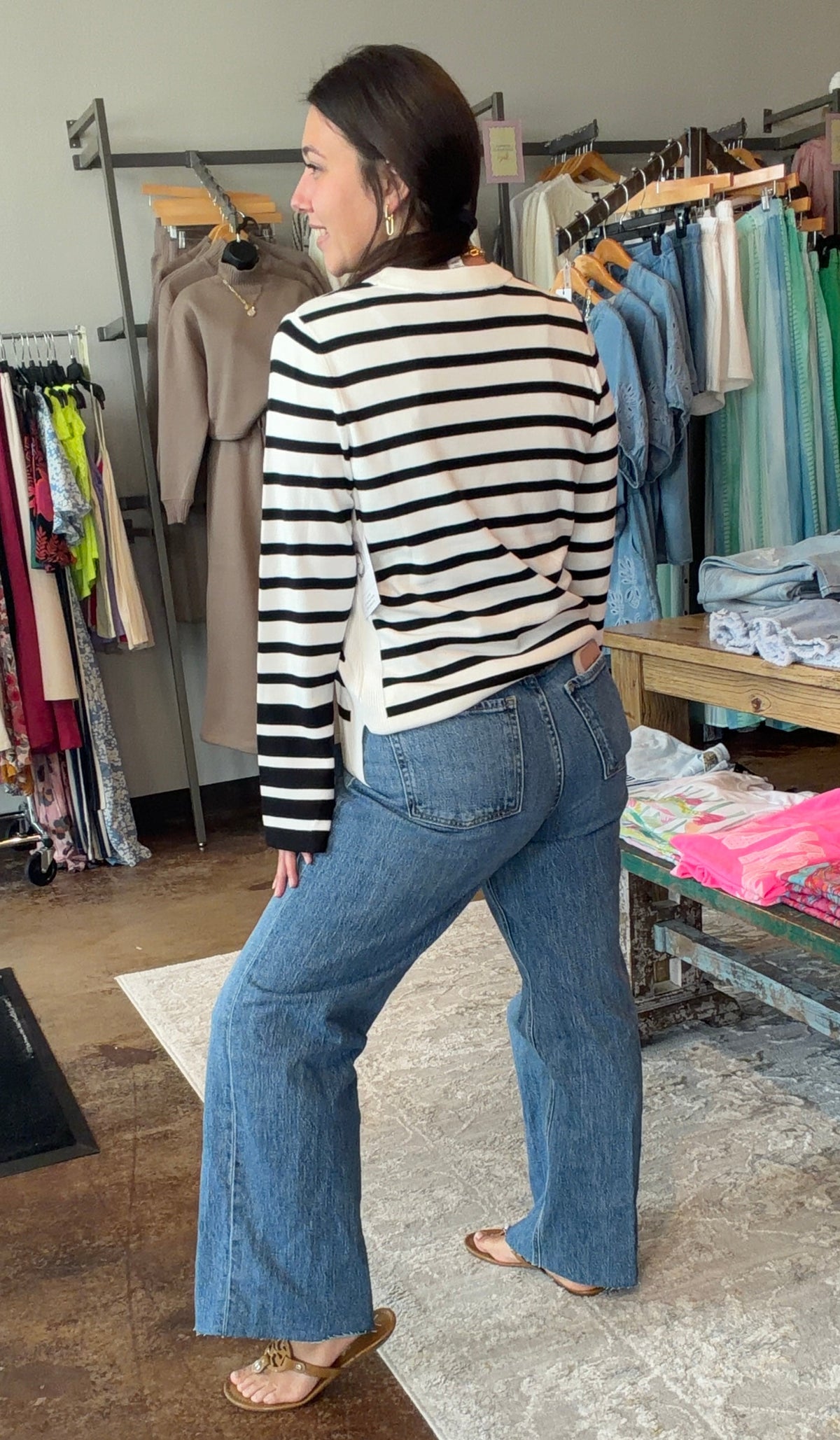 A woman wearing a black and white striped long-sleeve top and blue flare jeans stands inside a clothing boutique, looking to the side. Clothing racks, a table with folded garments, and a patterned rug are visible in the background.