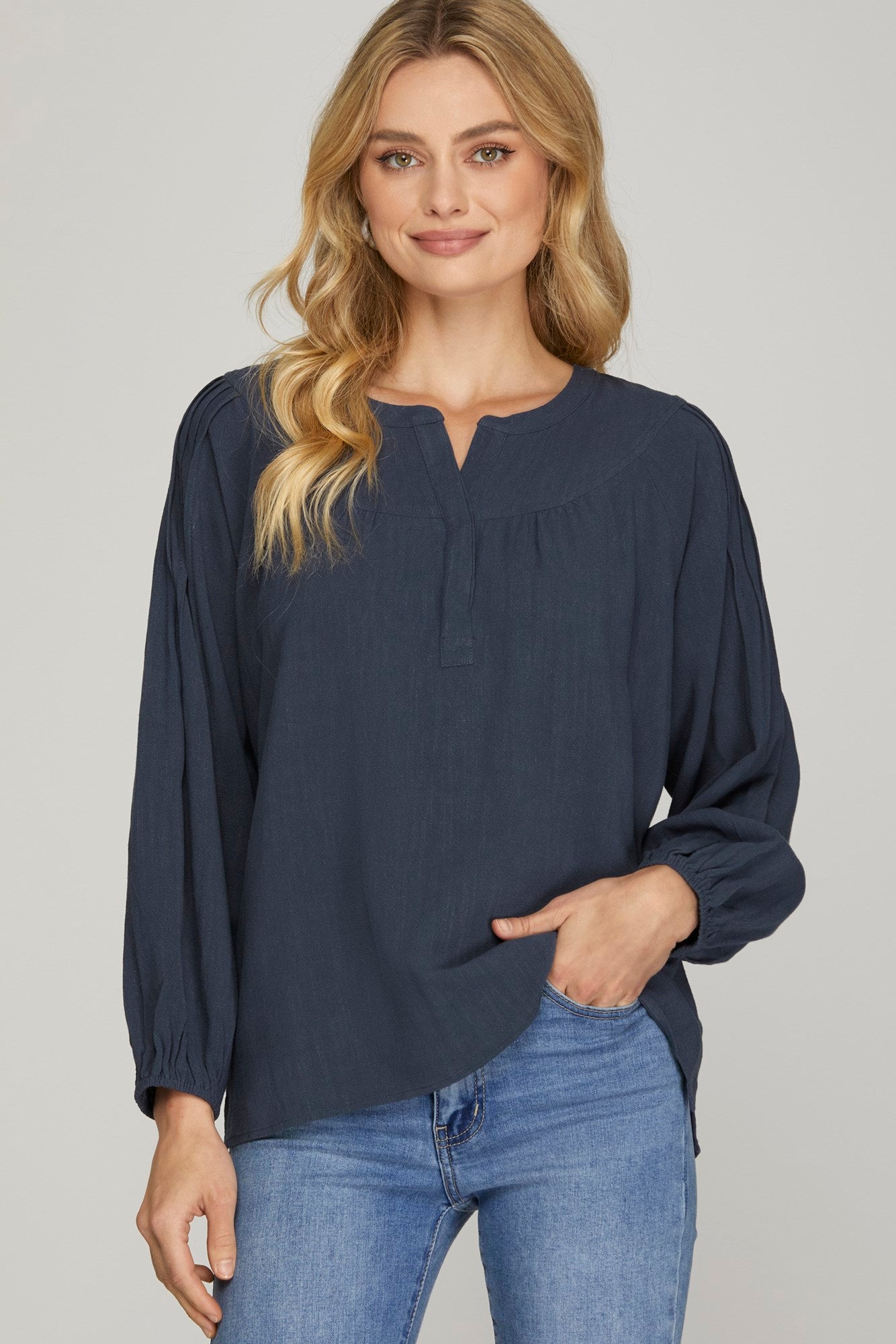 A woman wearing a navy blue long-sleeve blouse with a split neckline and blue jeans, standing against a light gray studio background.