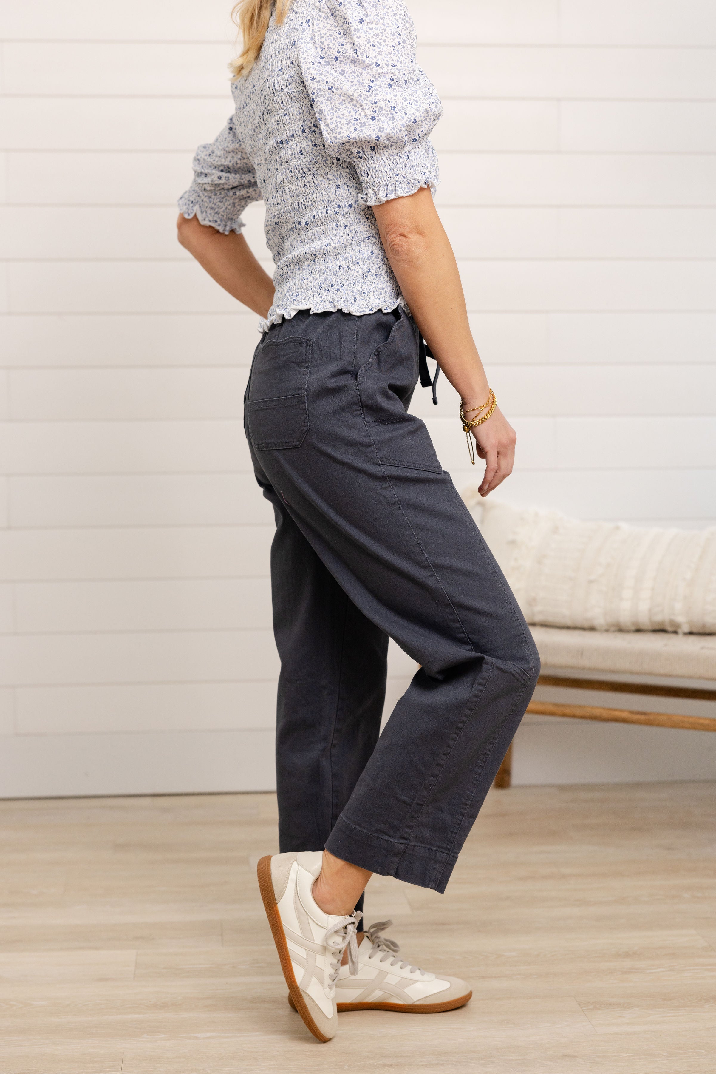 Person wearing a patterned top and dark pants with white sneakers in a minimal indoor setting.