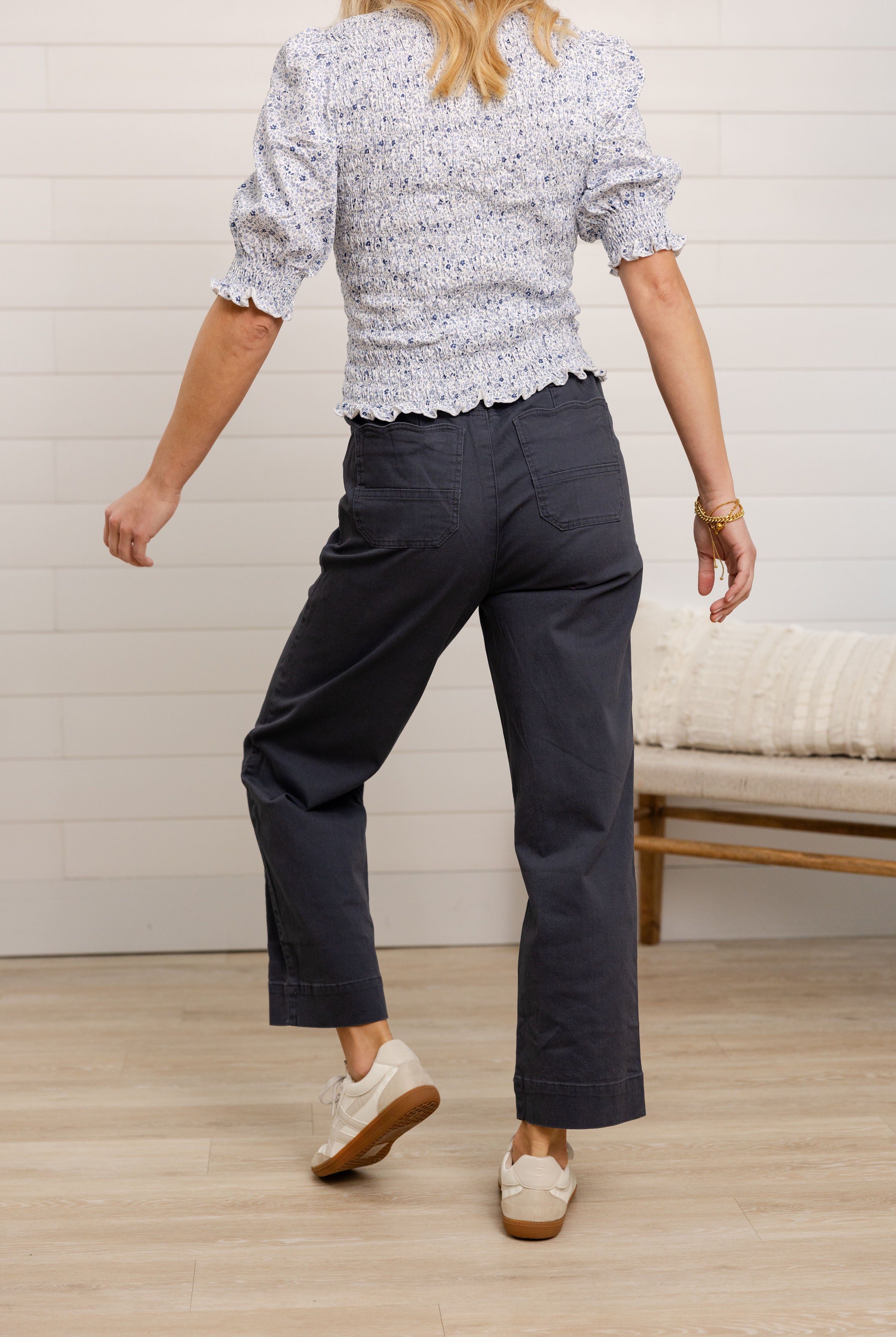 Person wearing a patterned top and dark pants in a room with a wooden floor and white wall.