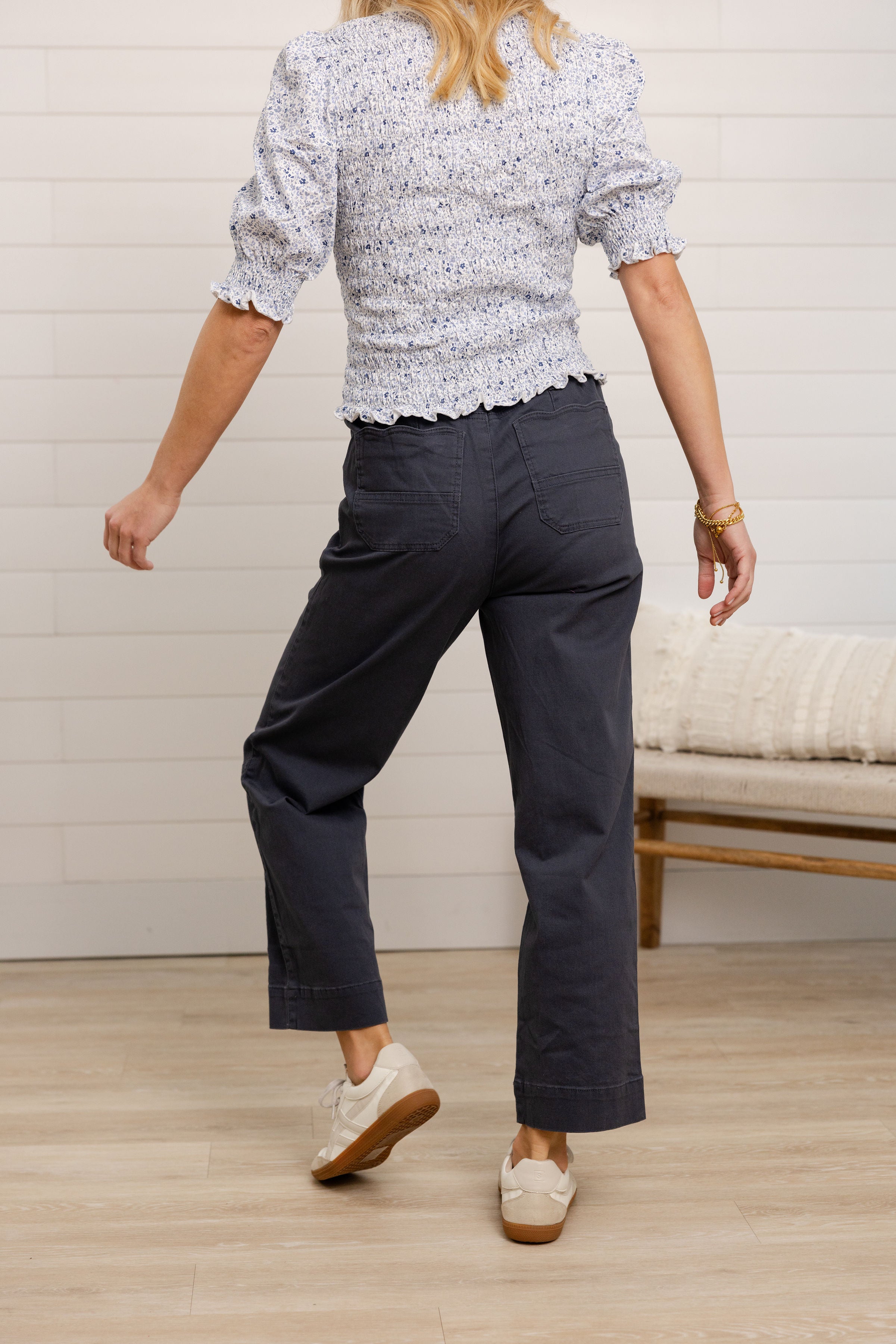 Person wearing a patterned top and dark pants in a room with a wooden floor and white wall.