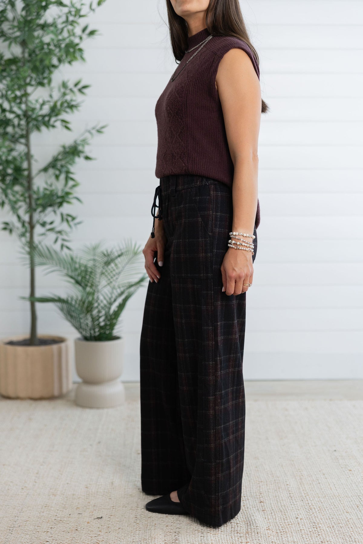 A woman wearing a sleeveless burgundy knit top and dark brown plaid wide-leg pants, standing in a bright room with potted plants and a white paneled wall.