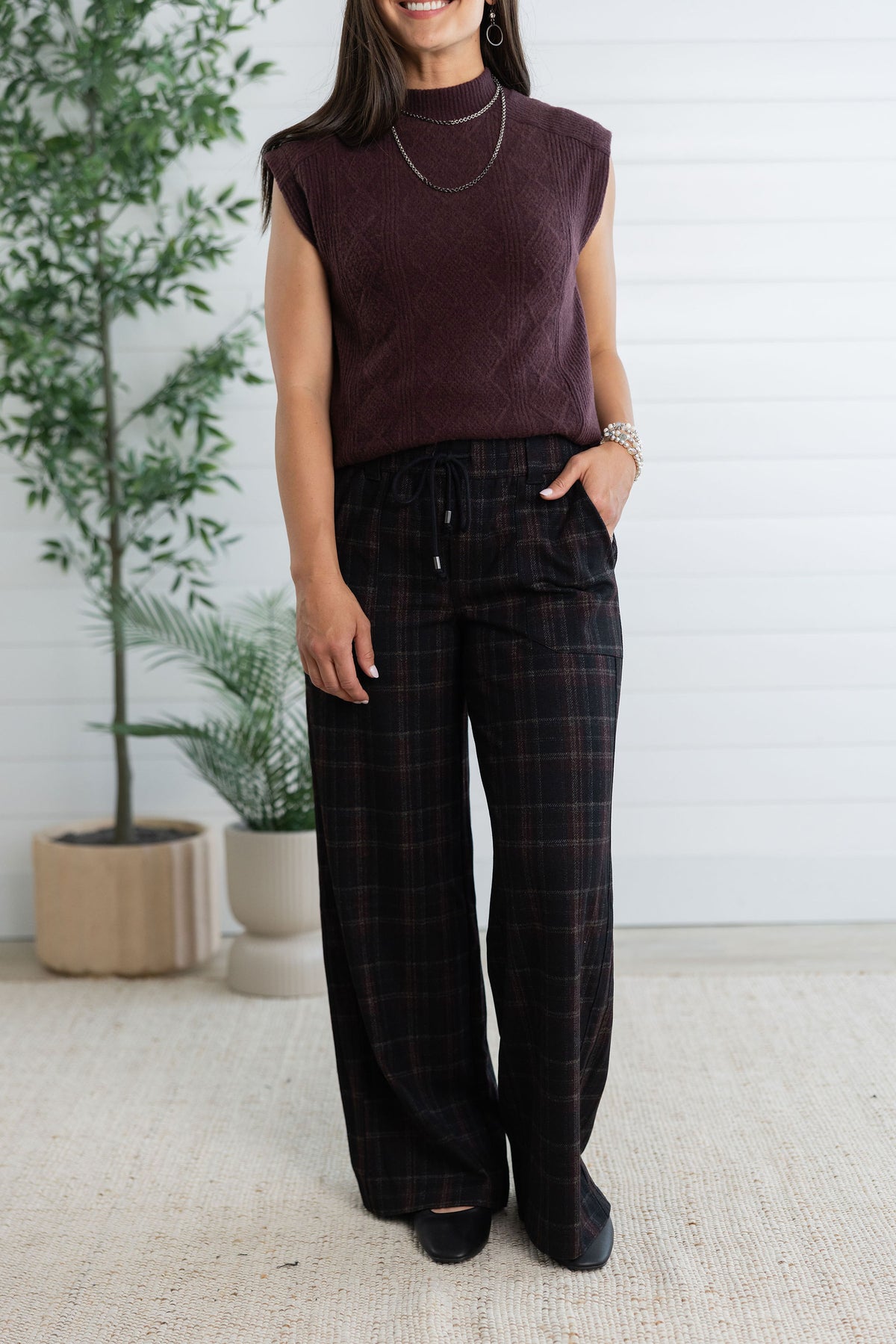 A woman wearing a sleeveless dark plum knit top and dark plaid wide-leg pants, standing in a bright indoor setting with potted plants and a white paneled wall.