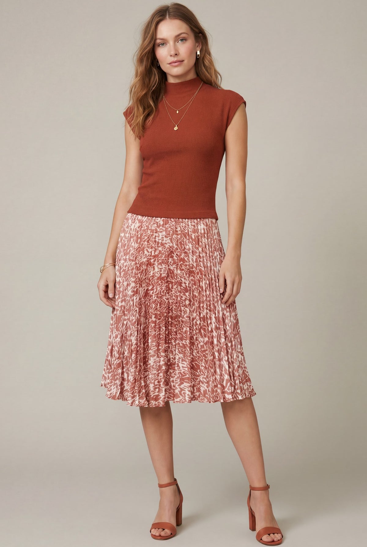 A woman modeling a rust-colored sleeveless mock-neck top paired with a pleated midi skirt in a pink and red abstract pattern, against a solid light gray studio background.