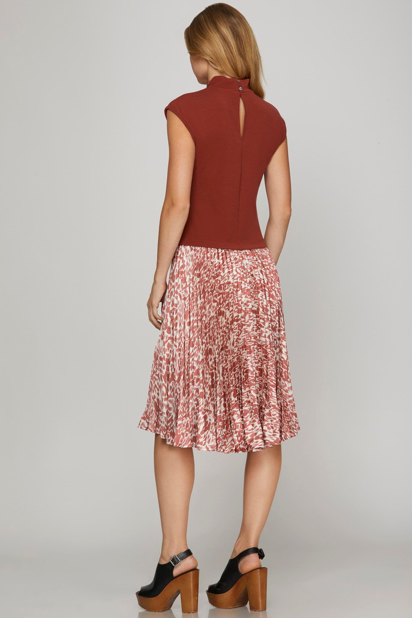 A woman shown from the back wearing a sleeveless rust-brown top and a pleated midi skirt with a pink, red, and white abstract pattern. She is wearing black platform sandals against a light gray studio background.