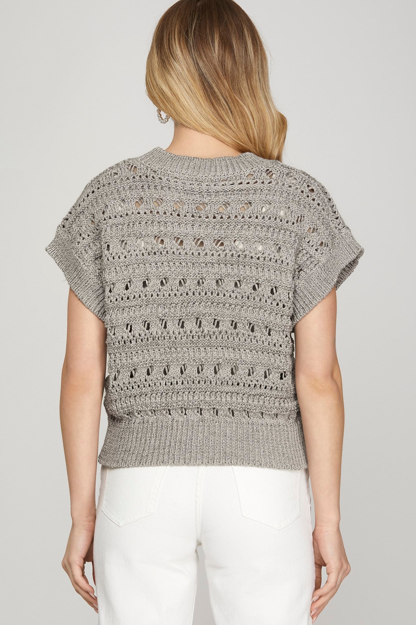 A woman shown from the back wearing a light gray short-sleeve knit top with textured openwork patterns and white pants, against a plain light gray studio background.