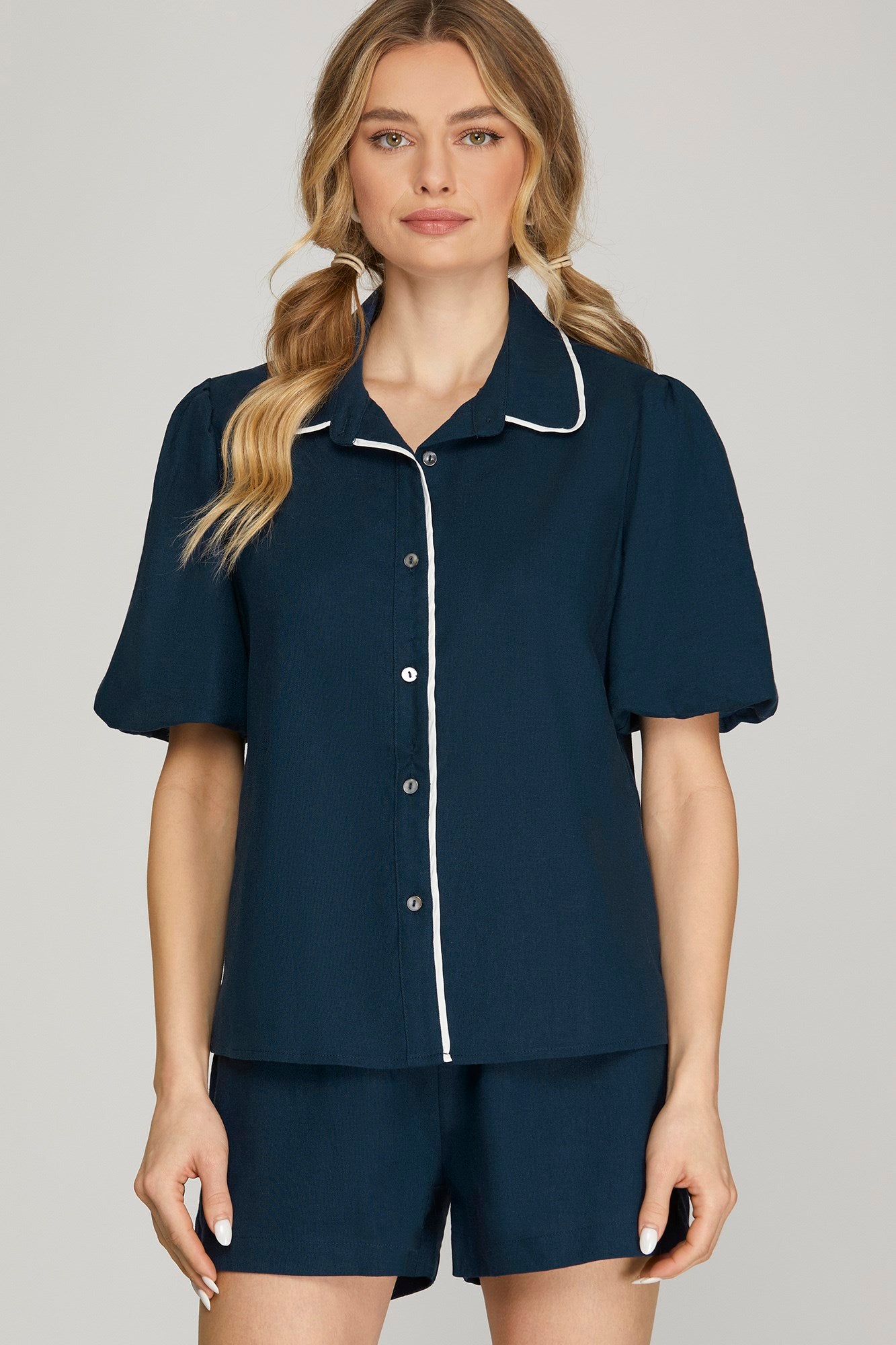 A woman modeling a navy blue two-piece pajama set with white piping details, consisting of a short-sleeve button-up shirt and matching shorts, against a solid light gray background.