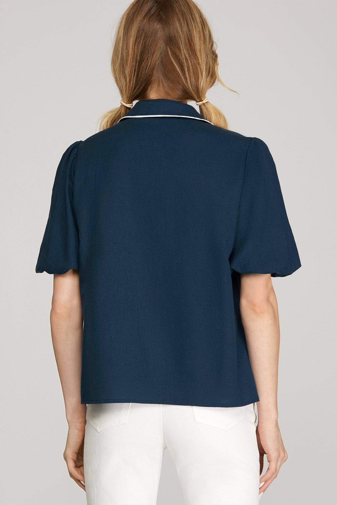 A woman shown from the back wearing a navy blue short-sleeve collared blouse with thin white piping trim and white pants, against a light gray studio background.