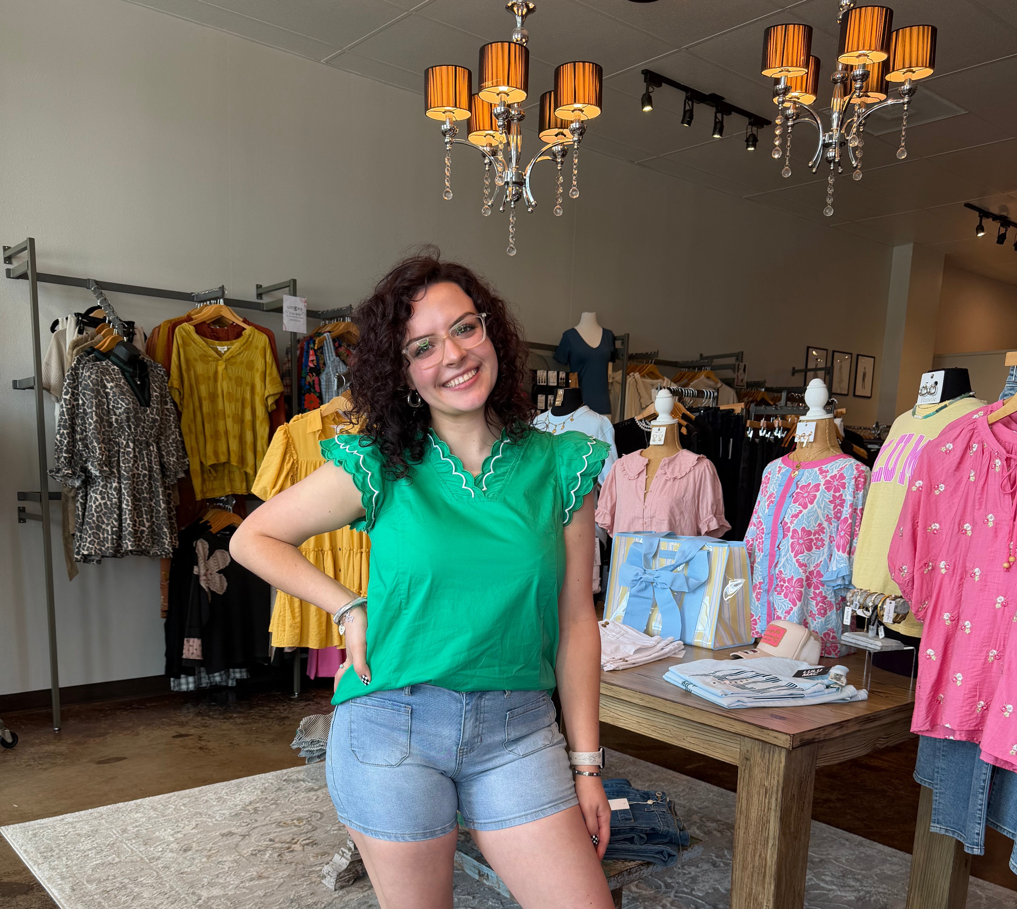 Woman in a green shirt and denim shorts standing in a store with clothing racks and displays.