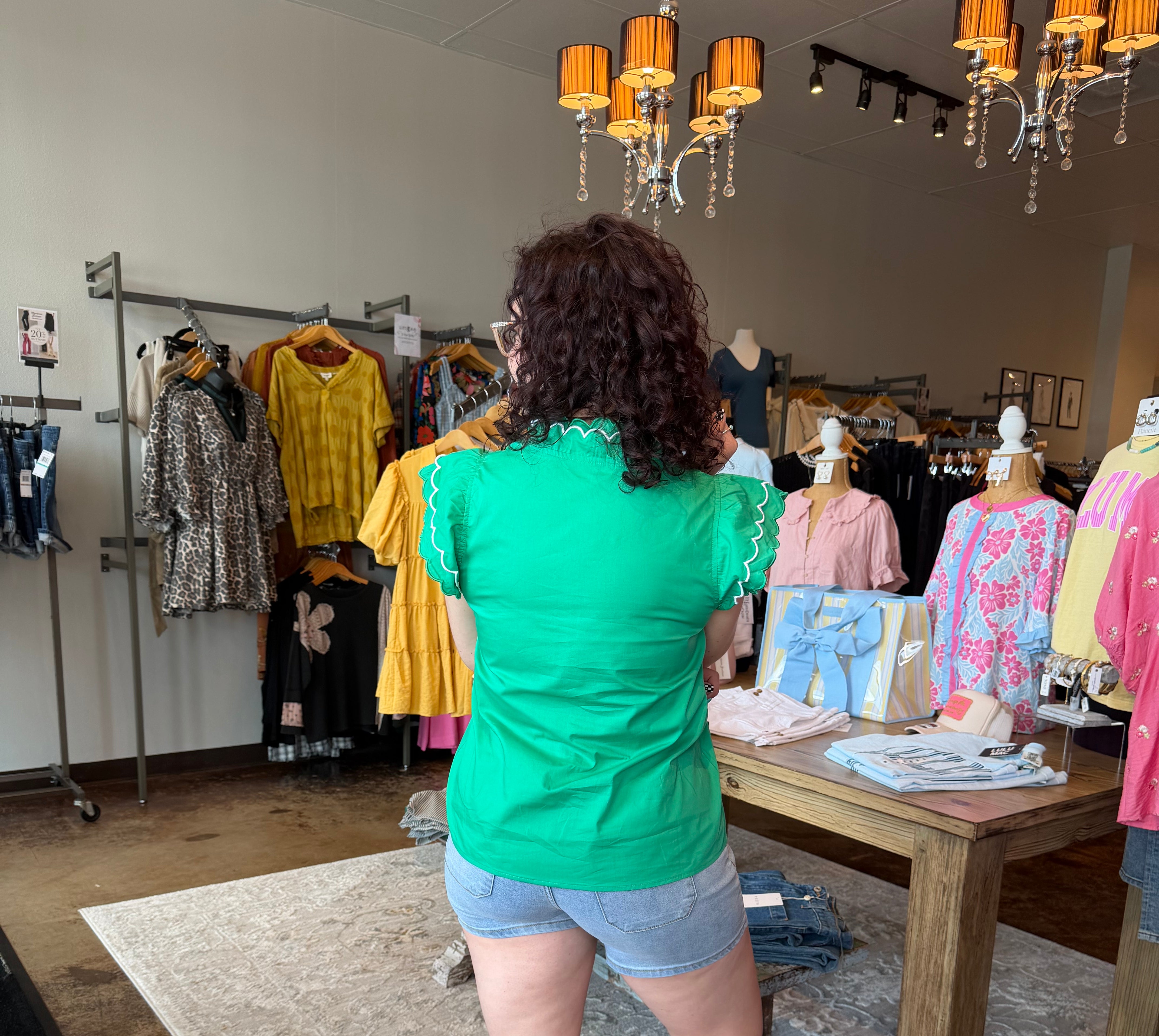 Woman in a green shirt and shorts standing in a clothing store with racks of clothes and a chandelier.