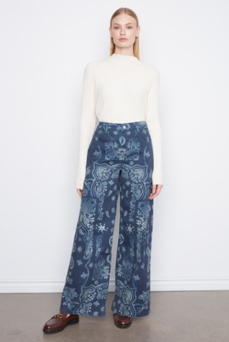 A model stands against a white wall wearing a cream ribbed mock-neck top and high-waisted, wide-leg trousers with a blue and white paisley pattern. She is wearing brown leather loafers.