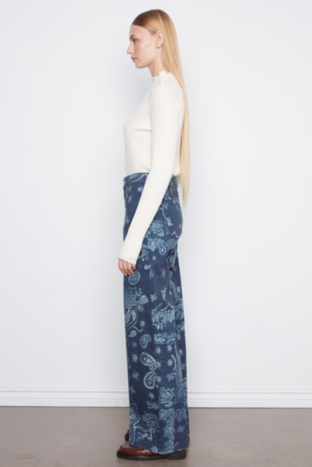 A woman stands in profile wearing a cream ribbed mock-neck long-sleeve top and high-waisted, wide-leg navy blue pants with a light blue paisley pattern, set against a plain white wall and light gray floor.