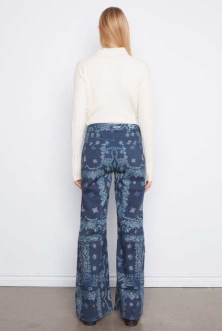 A woman viewed from behind wearing a cream ribbed mock-neck sweater and blue patterned wide-leg pants, standing against a plain white wall.