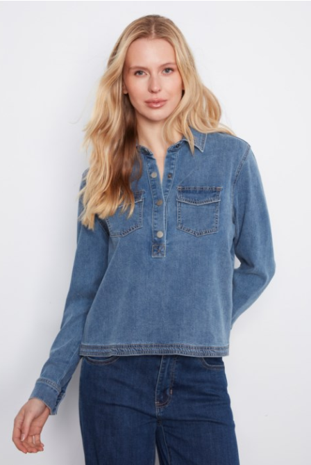 A blonde woman wearing a cropped, long-sleeve blue denim shirt-jacket with a buttoned henley-style placket and a chest pocket, paired with dark blue jeans, against a plain white studio background.