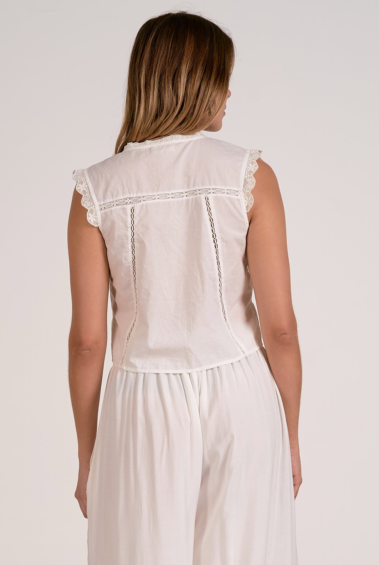 Woman wearing a white sleeveless top and pants with lace details on a plain background