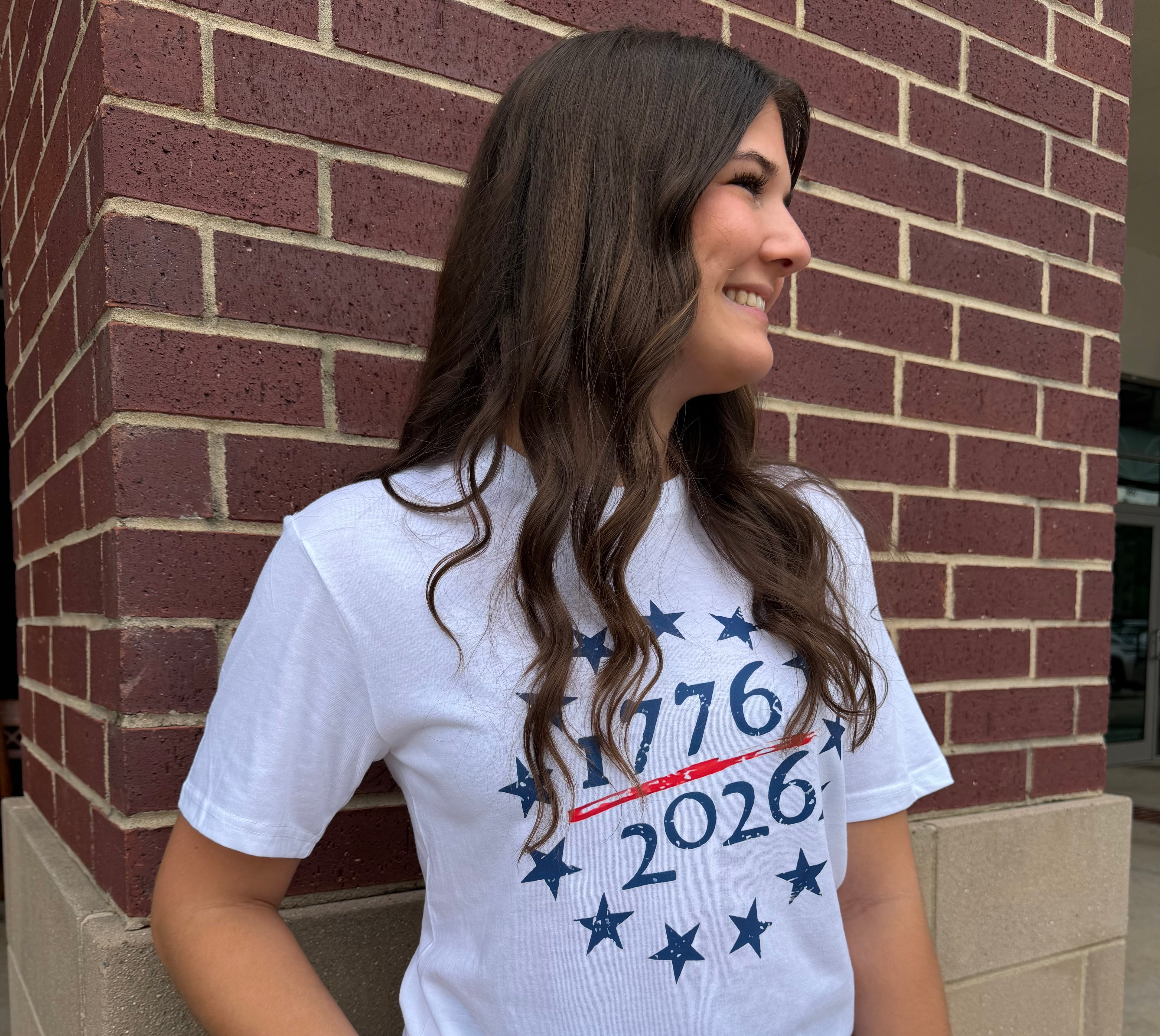 Woman wearing a white t-shirt with '1776 2026' design, standing against a brick wall.
