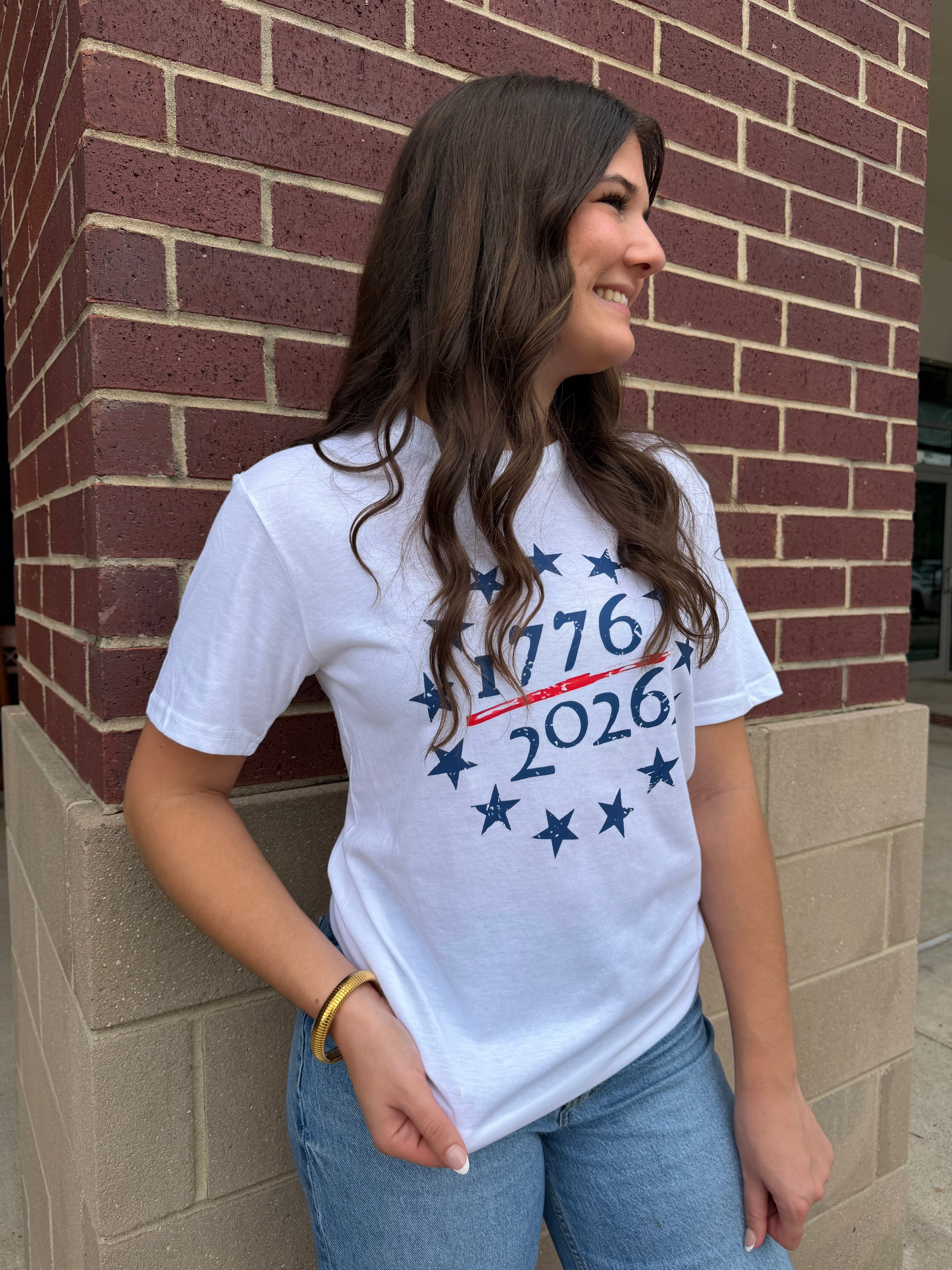 Woman wearing a white t-shirt with '1776 2026' design, standing against a brick wall.