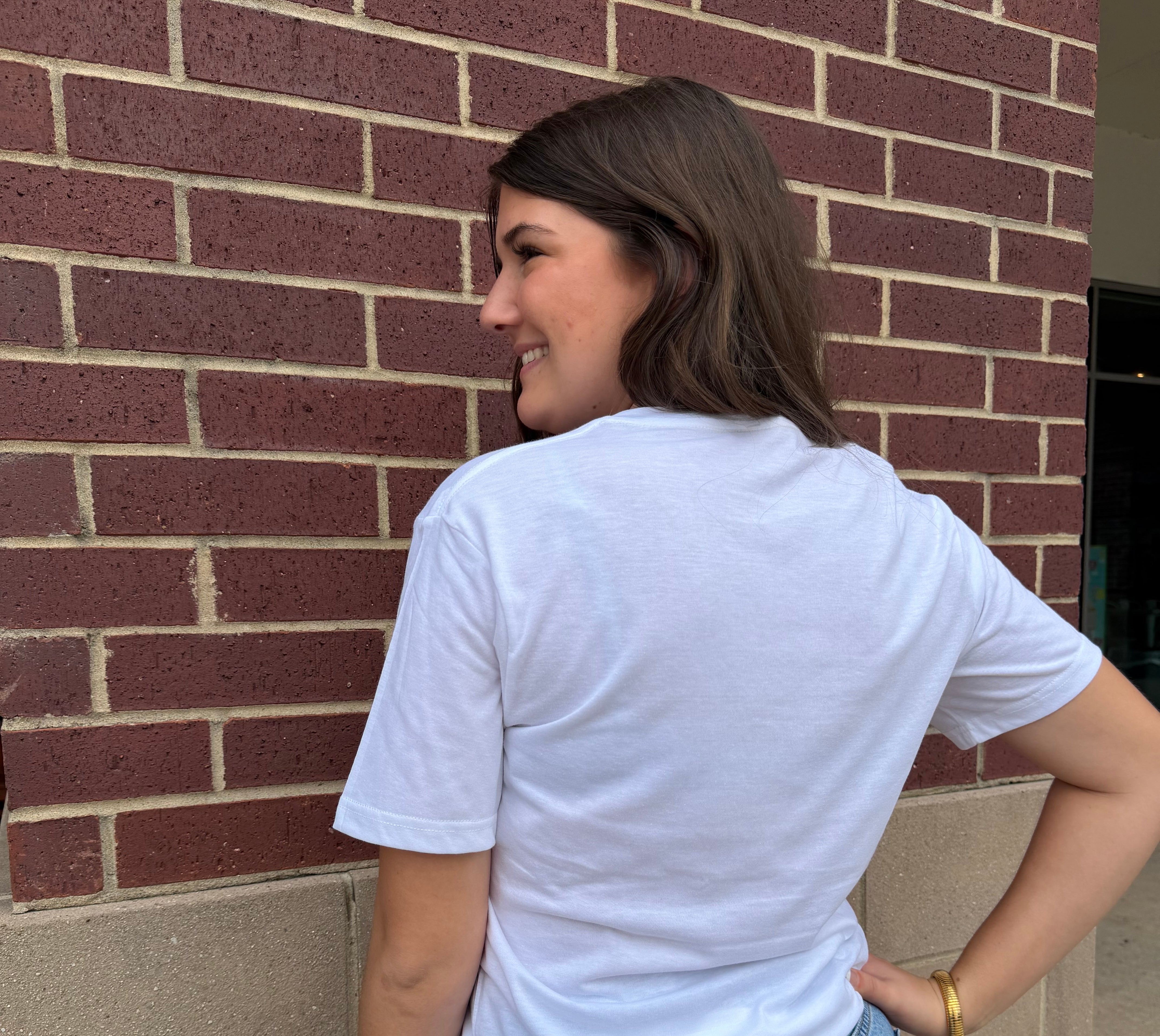 Person wearing a white t-shirt and blue jeans standing against a brick wall.