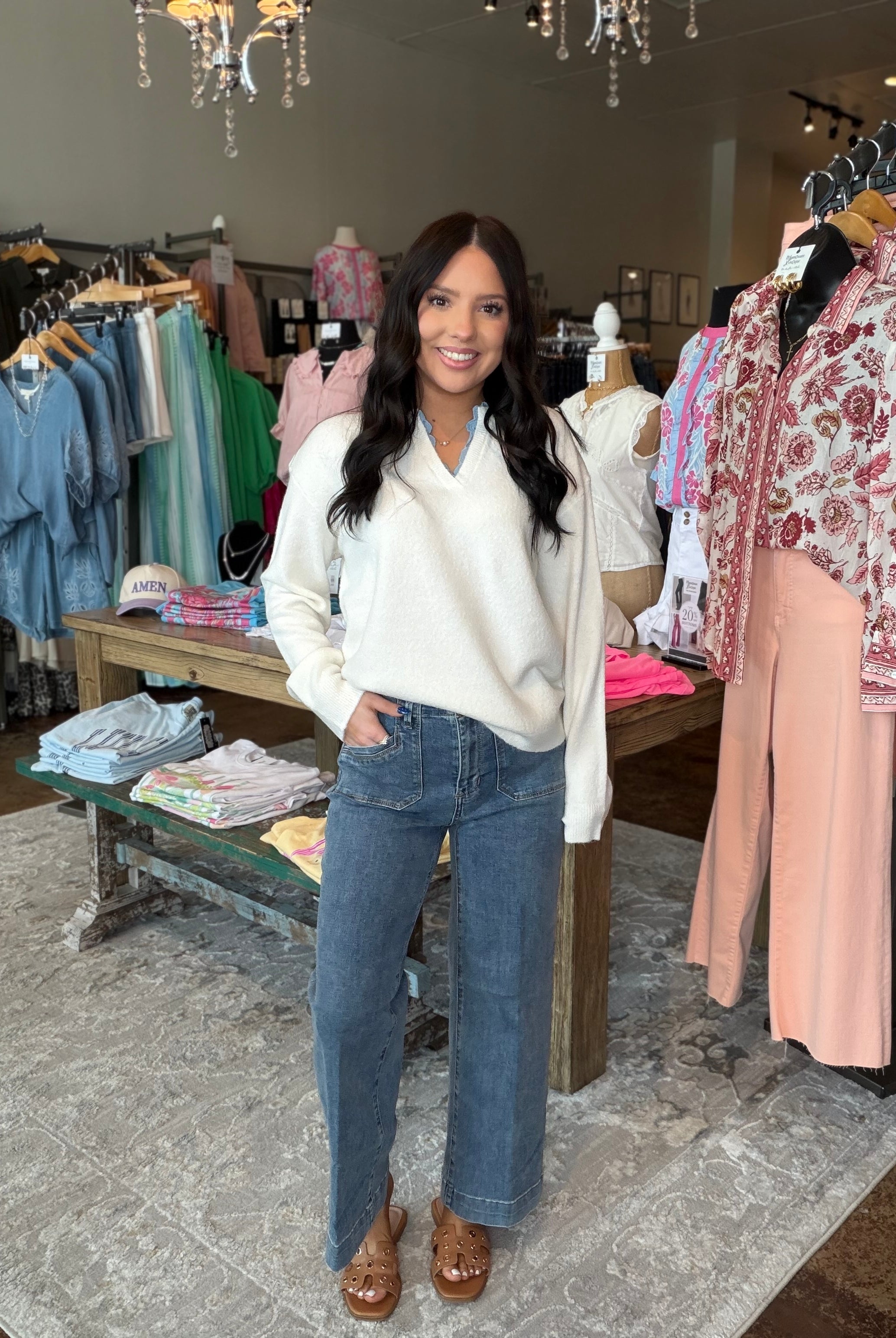 A woman stands in a clothing boutique wearing a cream long-sleeve V-neck sweater, blue wide-leg jeans, and brown leather wedge sandals. Clothing racks and mannequins are visible in the background.