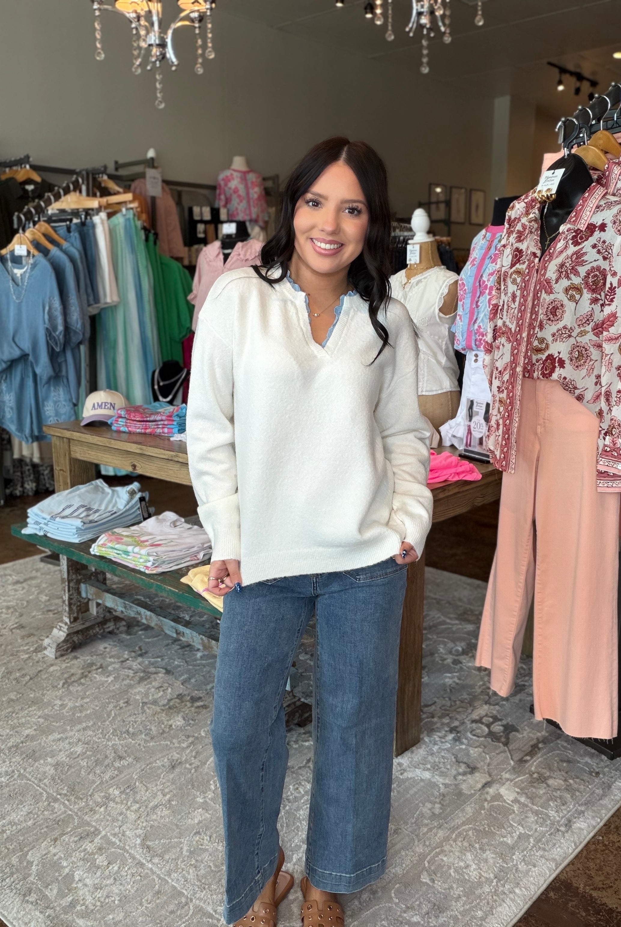 A woman stands in a clothing boutique wearing a cream V-neck knit sweater and wide-leg blue jeans, with racks of apparel and store fixtures visible around her.